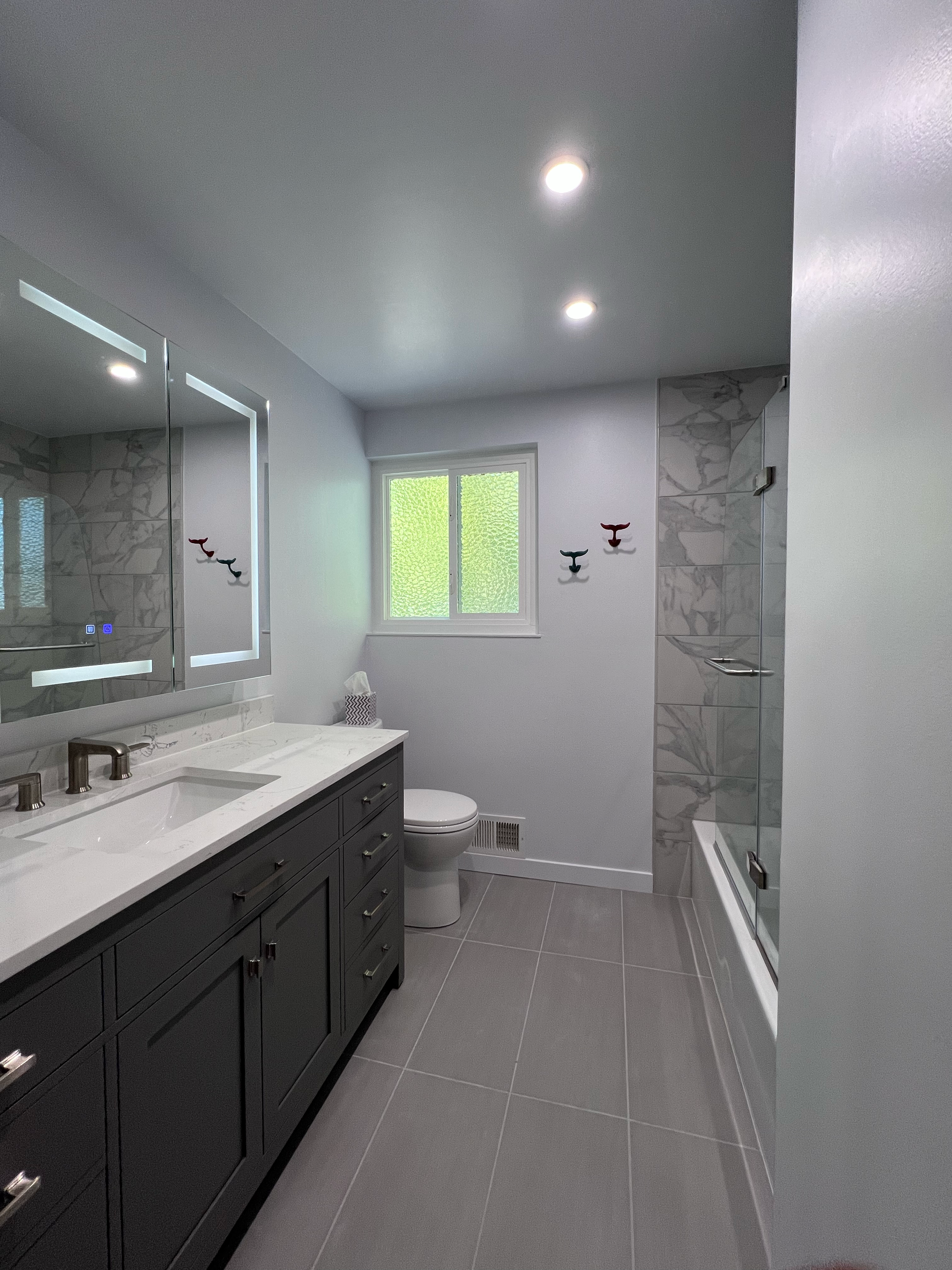 Modern bathroom with gray vanity, white countertop, wall-mounted mirror with lights, toilet, and shower with glass door, gray tiled floor, and wall decorated with whale-shaped hooks.