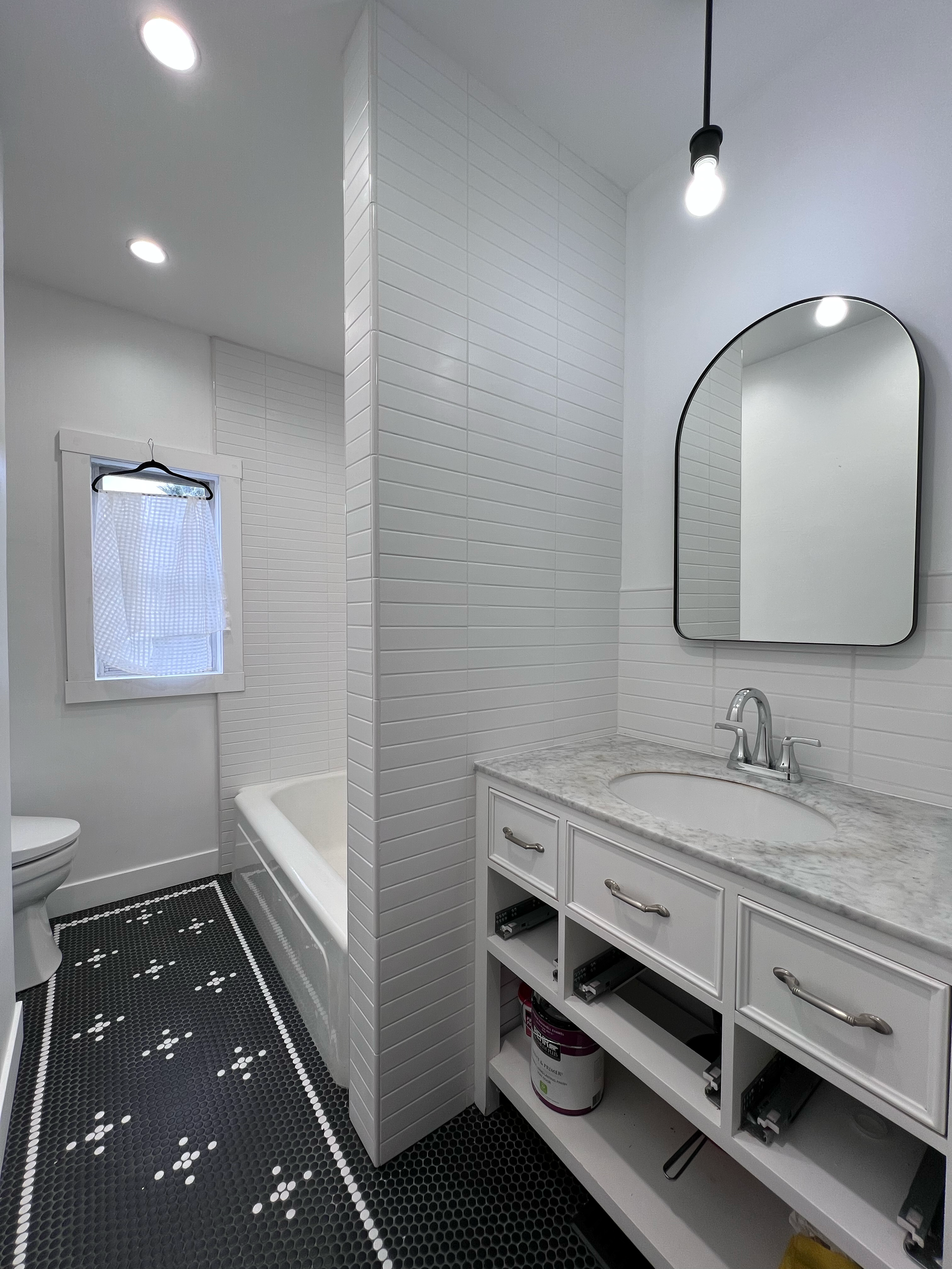 A bathroom with black hexagonal tile flooring featuring white dotted accents and a white vanity with a marble countertop, an oval mirror, and a modern faucet. There is a small window with a checkered curtain, a bathtub, and ceiling lights.