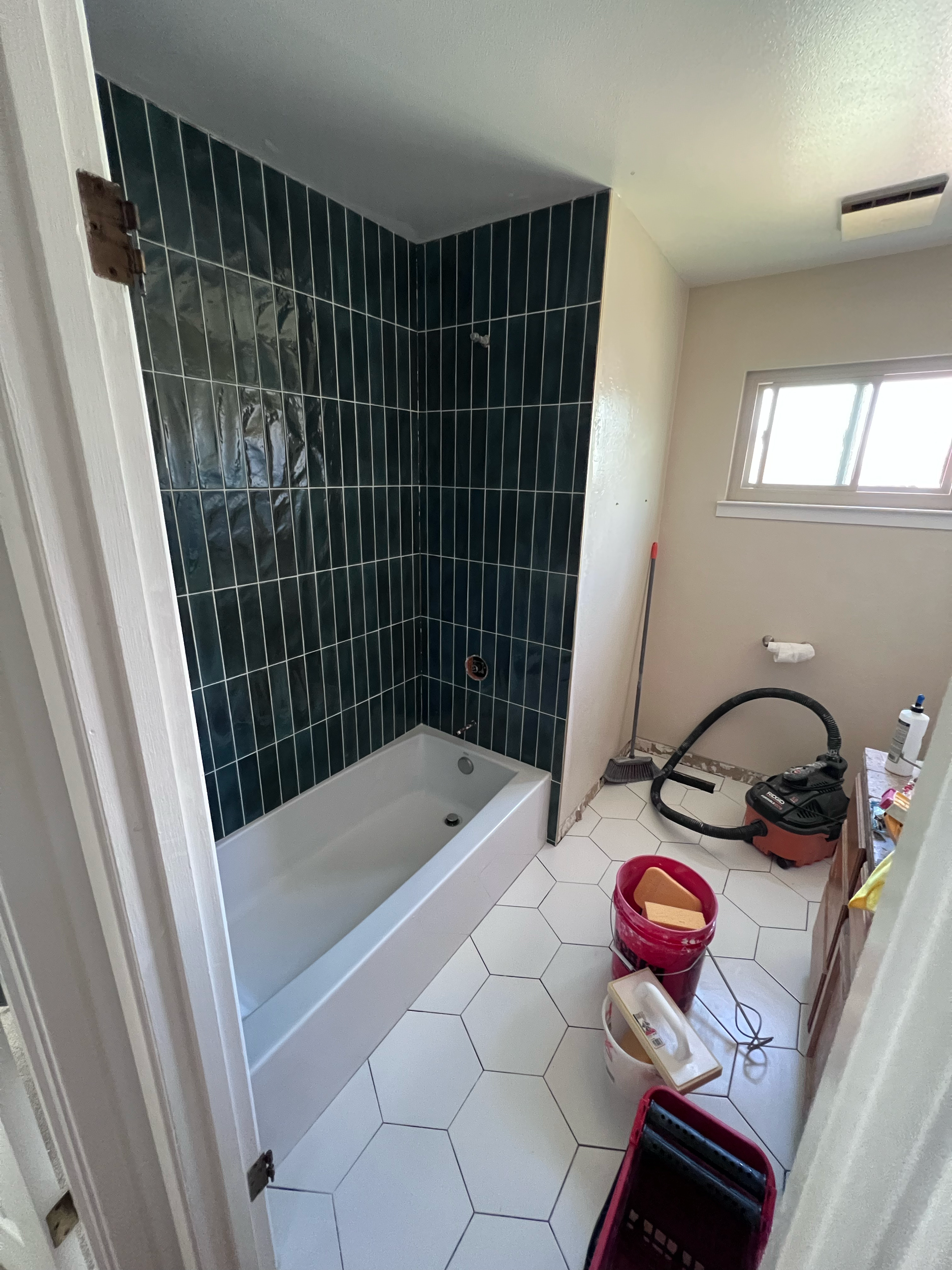Bathroom under renovation with black tiled shower area, white hexagonal floor tiles, and construction tools and supplies scattered around.