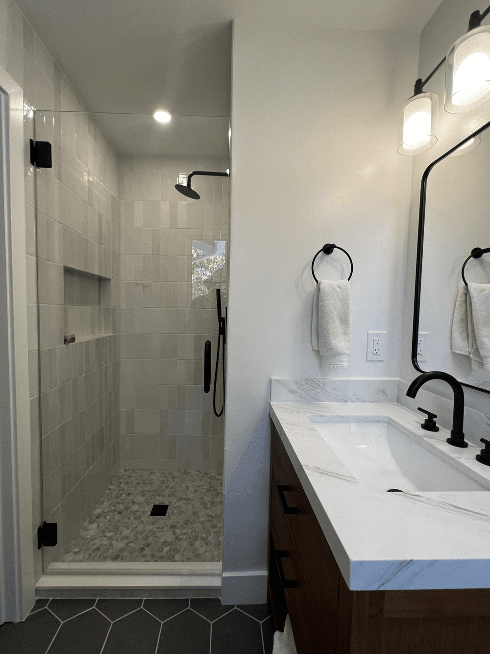 Modern bathroom remodel with a glass-enclosed shower, white tiled walls, marble countertop, dark wood vanity, black fixtures and a mirror with sconce lighting