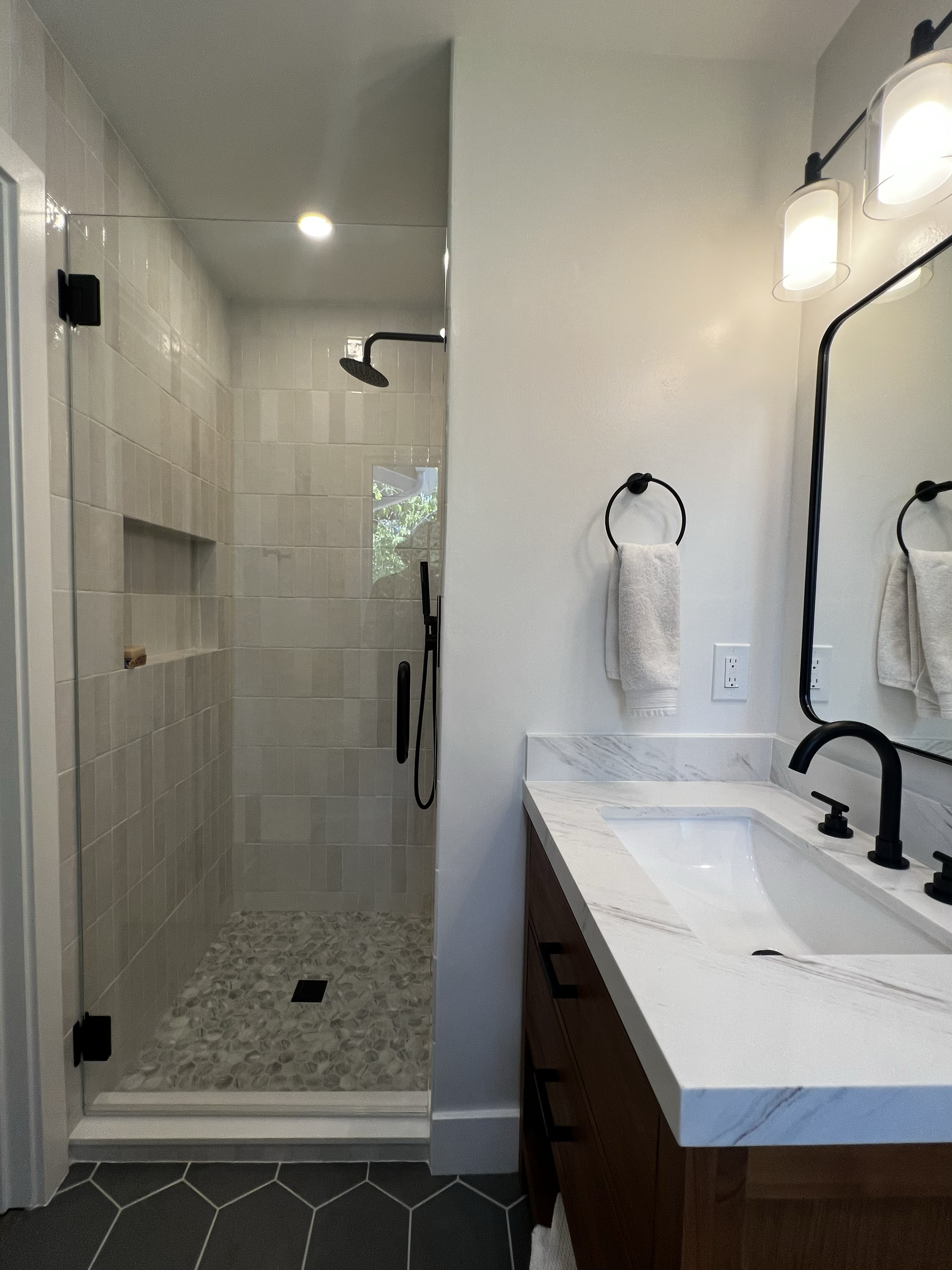 Modern bathroom remodel with a glass-enclosed shower, white tiled walls, marble countertop, dark wood vanity, black fixtures and a mirror with sconce lighting