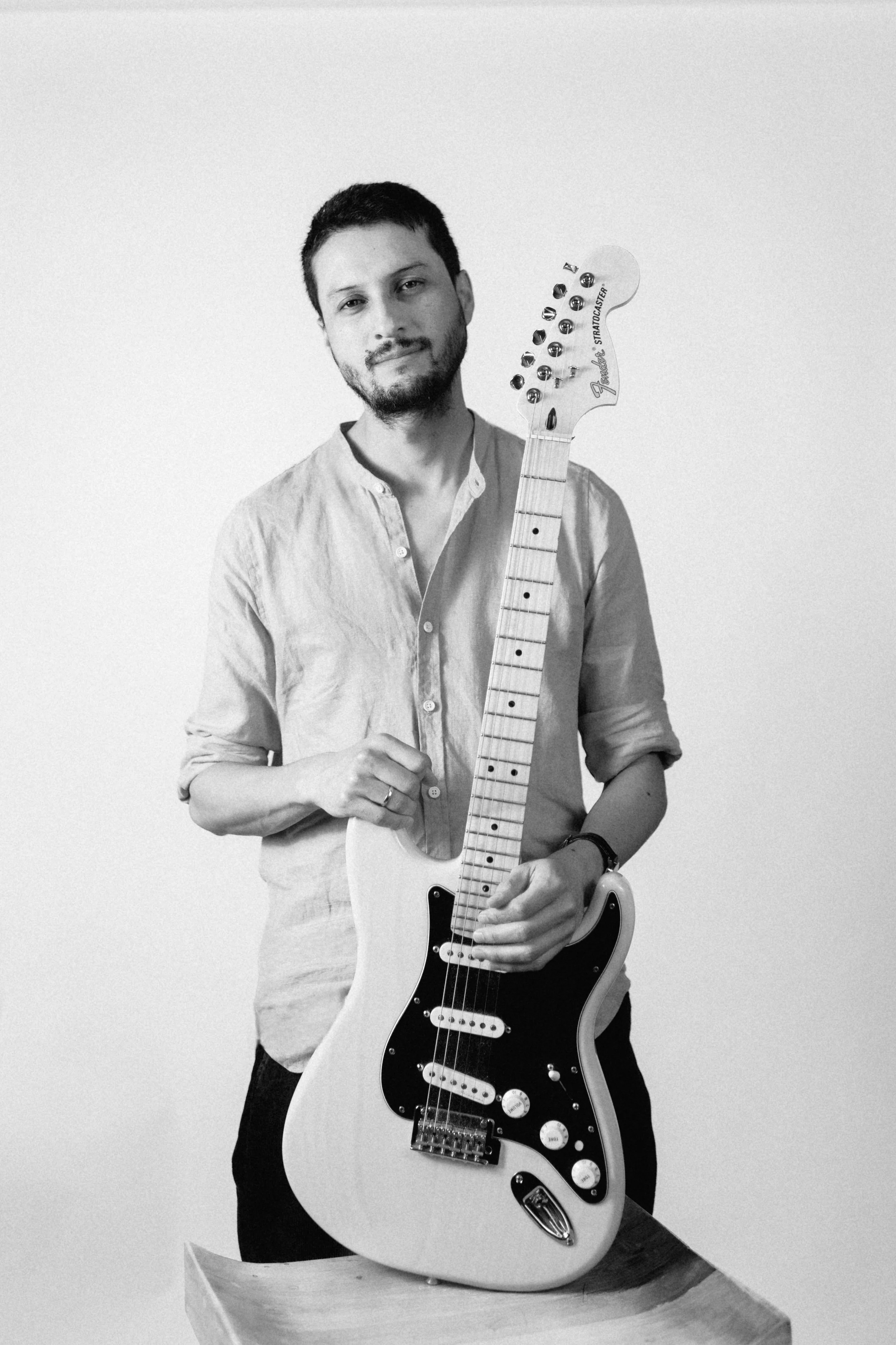 Un hombre con barba y camisas de manga larga sosteniendo una guitarra eléctrica Fender Stratocaster en un fondo blanco.