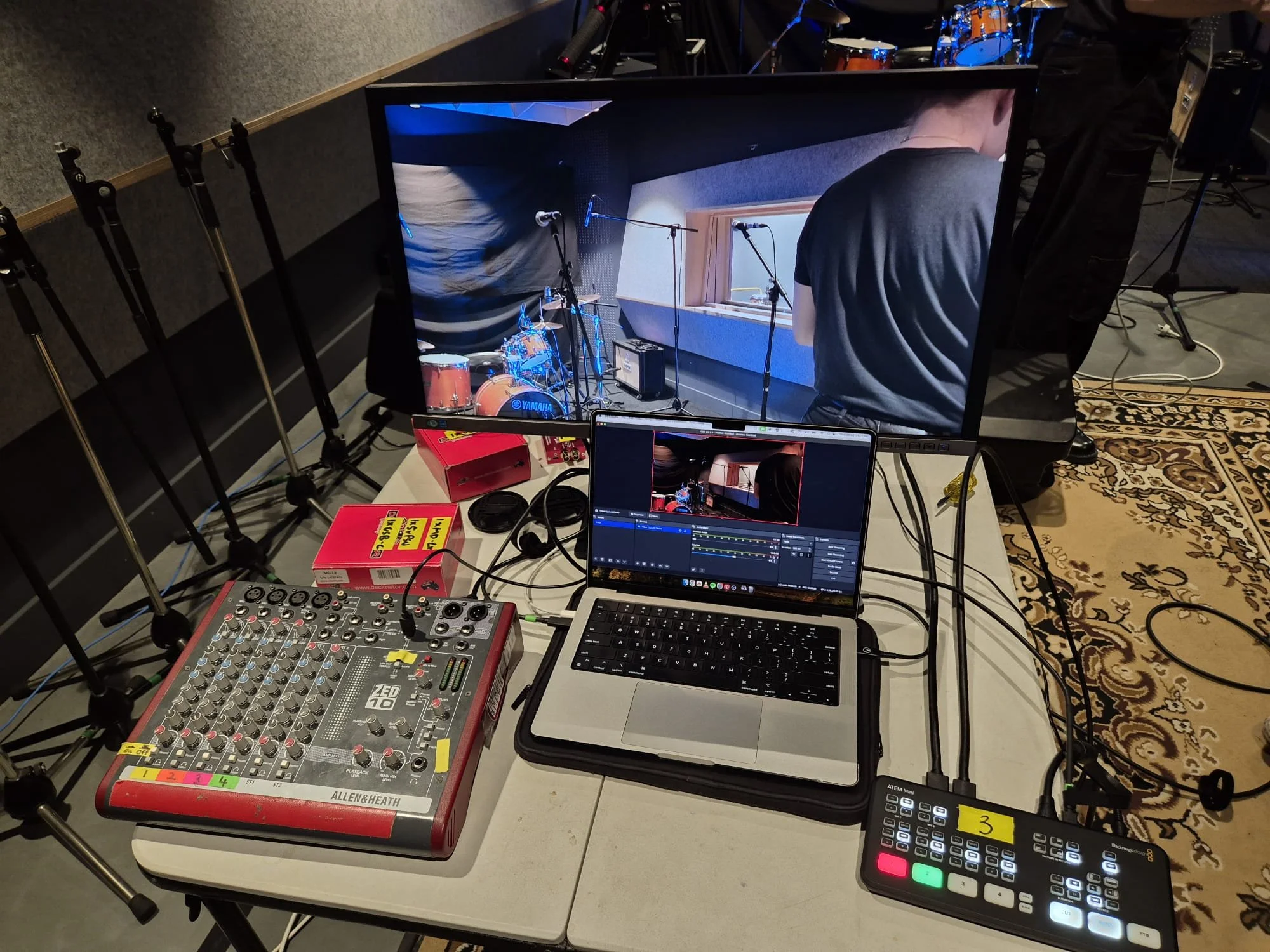 A live music recording setup with a laptop, audio mixer, and video monitor showing a band rehearsal in a music studio.
