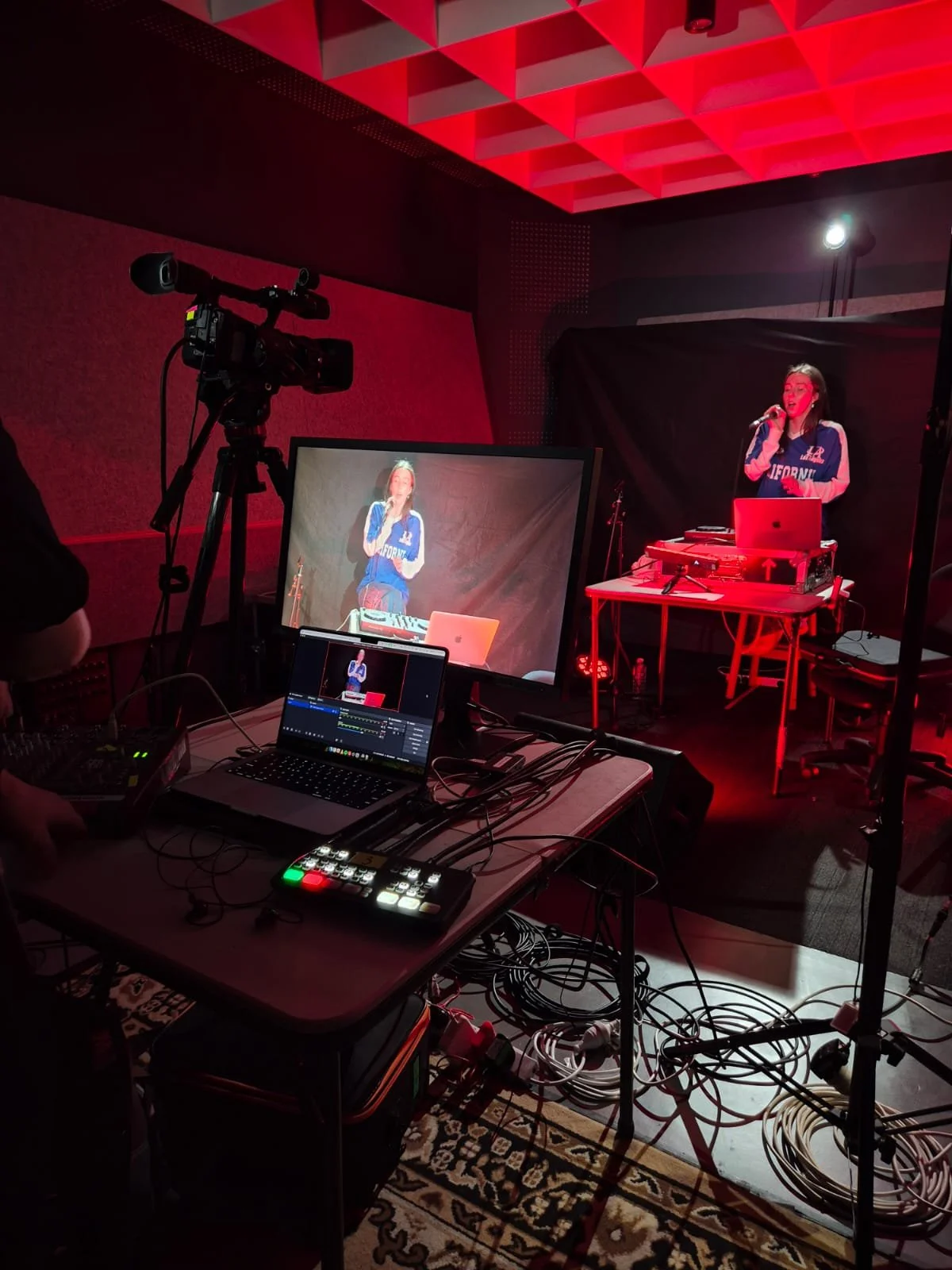 A woman stands on a small stage with a microphone, surrounded by audio-visual equipment, in a dimly lit room with red lighting.