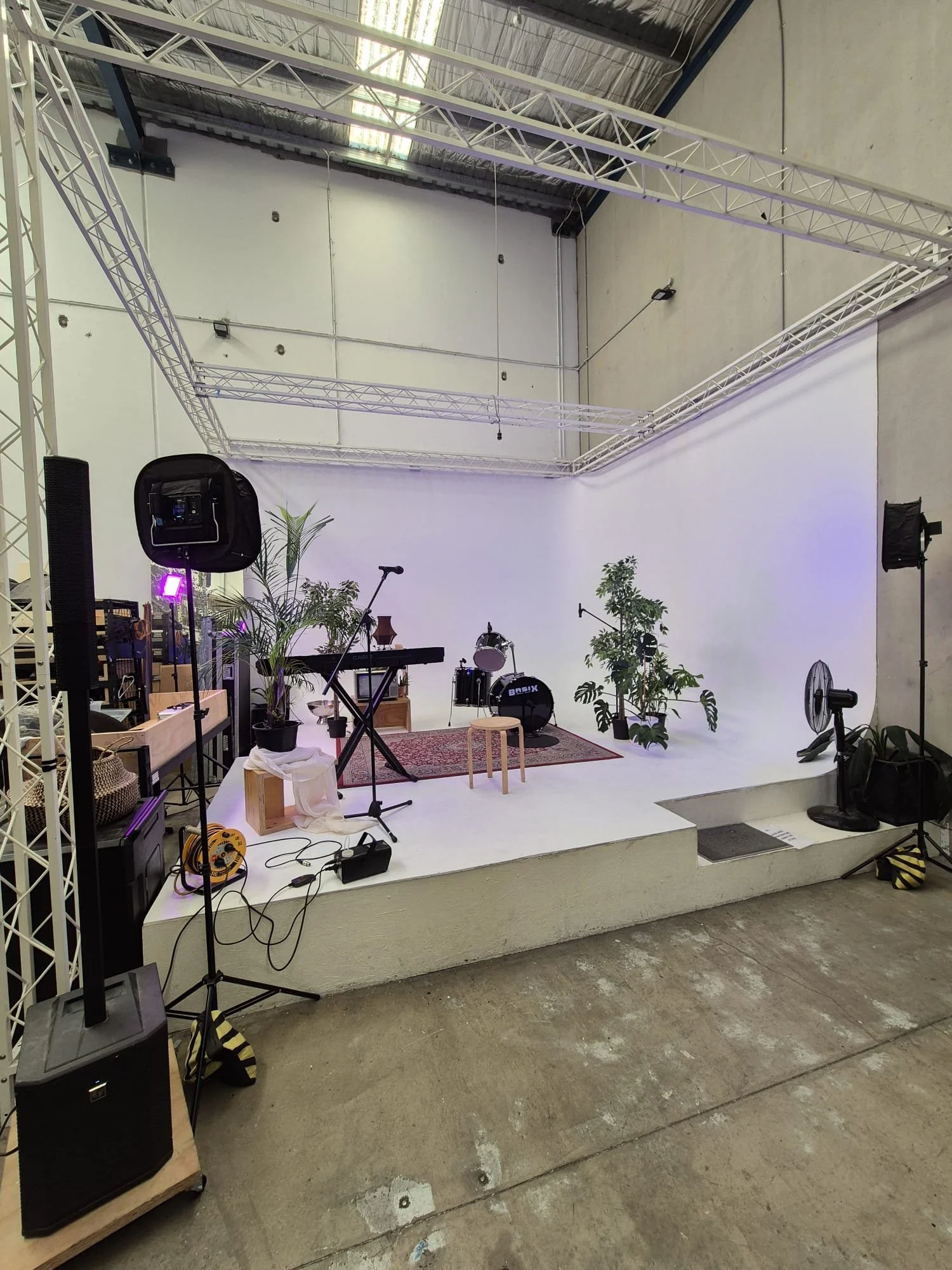 Empty stage setup with musical instruments, microphones, and plants, surrounded by professional lighting and sound equipment in a studio or warehouse space.