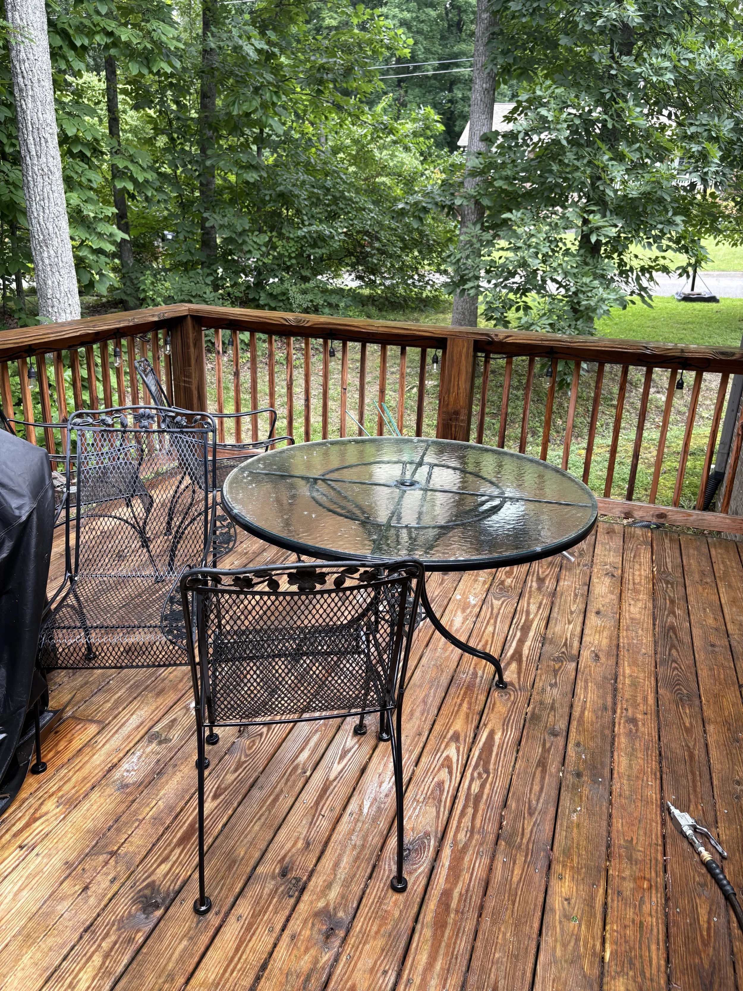 A wooden outdoor deck with a round glass-top table, four black metal chairs, and a black covered grill. There are trees and grassy yard in the background.
