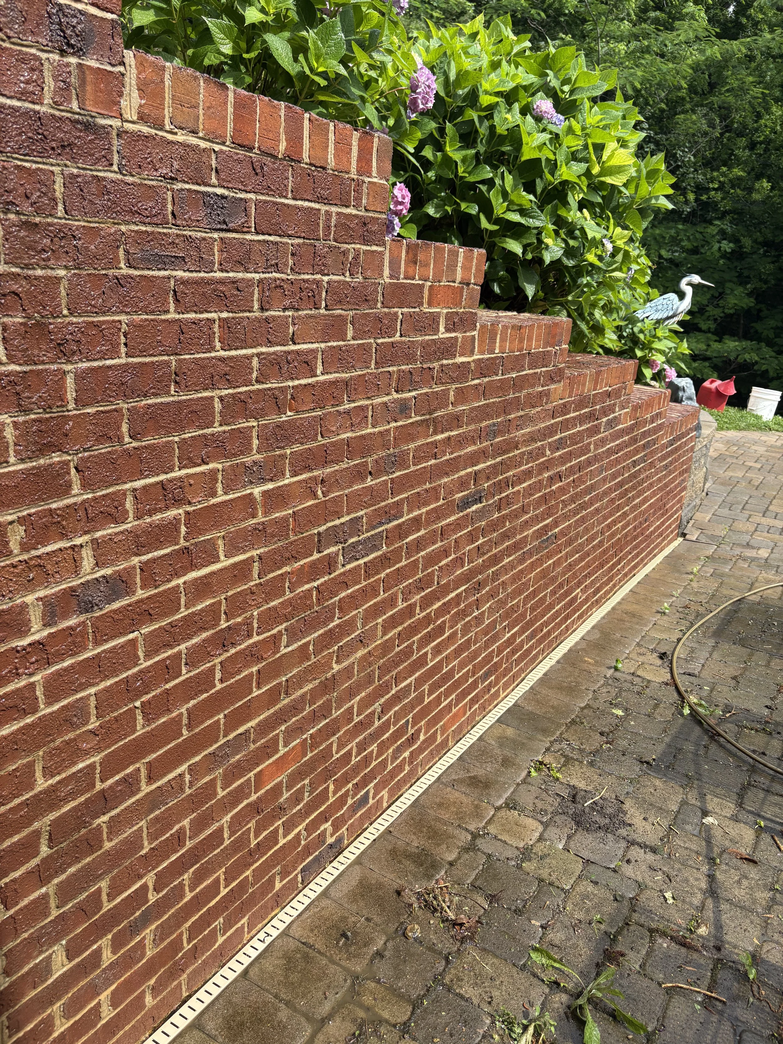 A red brick wall with a stepped top, next to a paved patio with some scattered leaves and a garden hose. Behind the wall, green bushes with purple flowers and a bird sculpture are visible.