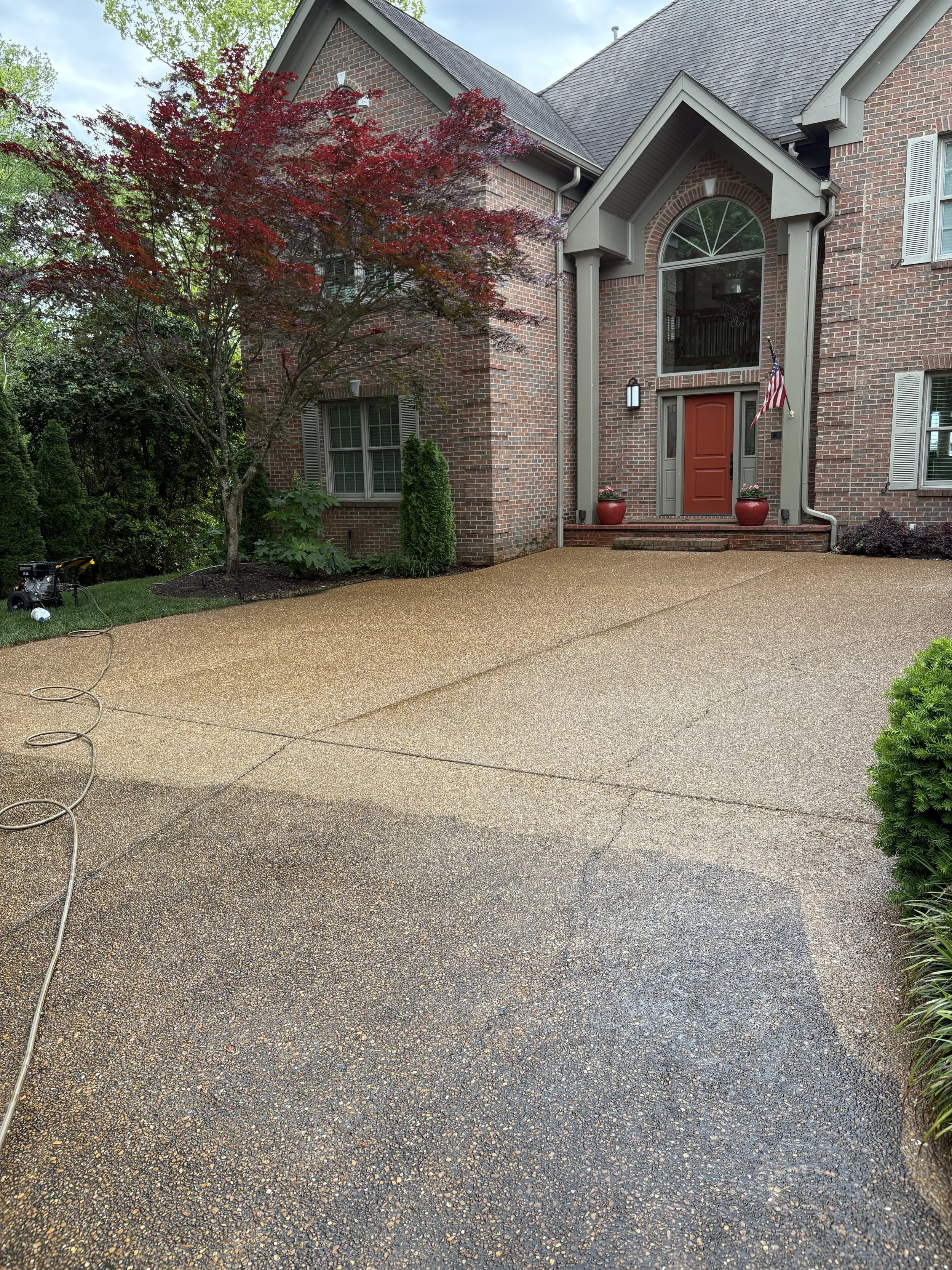 A house with brick exterior, a large front porch with steps, potted plants, an American flag, and a large window above the front door. There are trees and shrubs around the house, and a wet concrete driveway with a hose and pressure washer on it.
