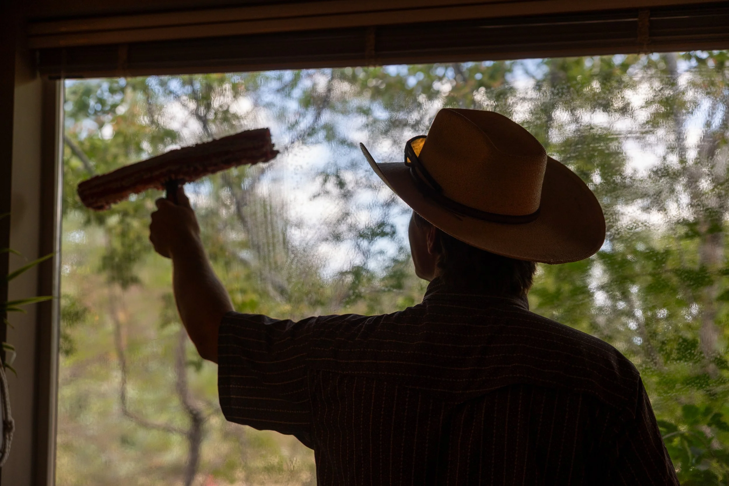 A man wearing a straw hat is cleaning a window with a squeegee, viewed from inside a house with a natural landscape outside.