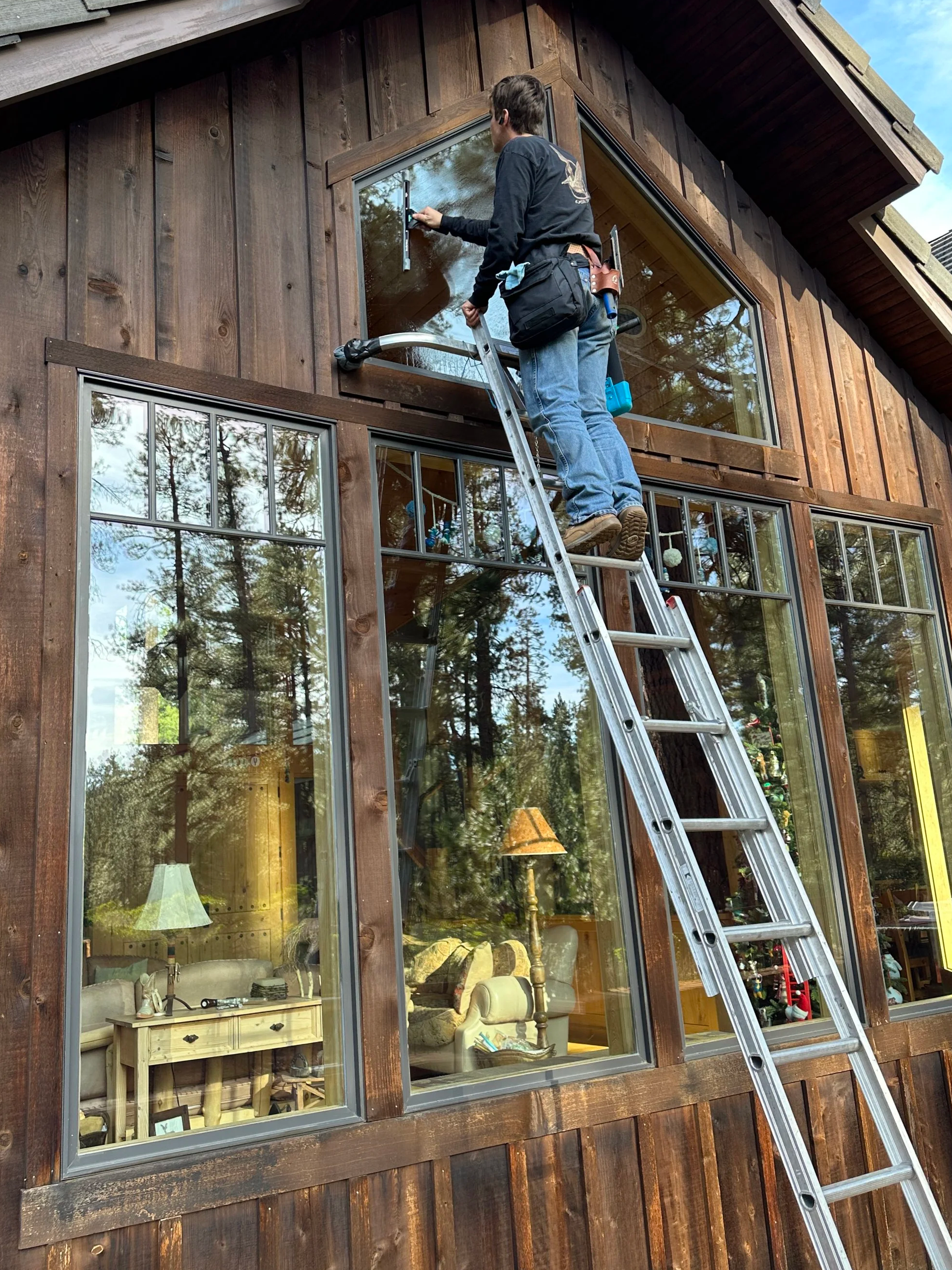 A man is standing on a ladder cleaning or installing a large upper window on a wooden house exterior.