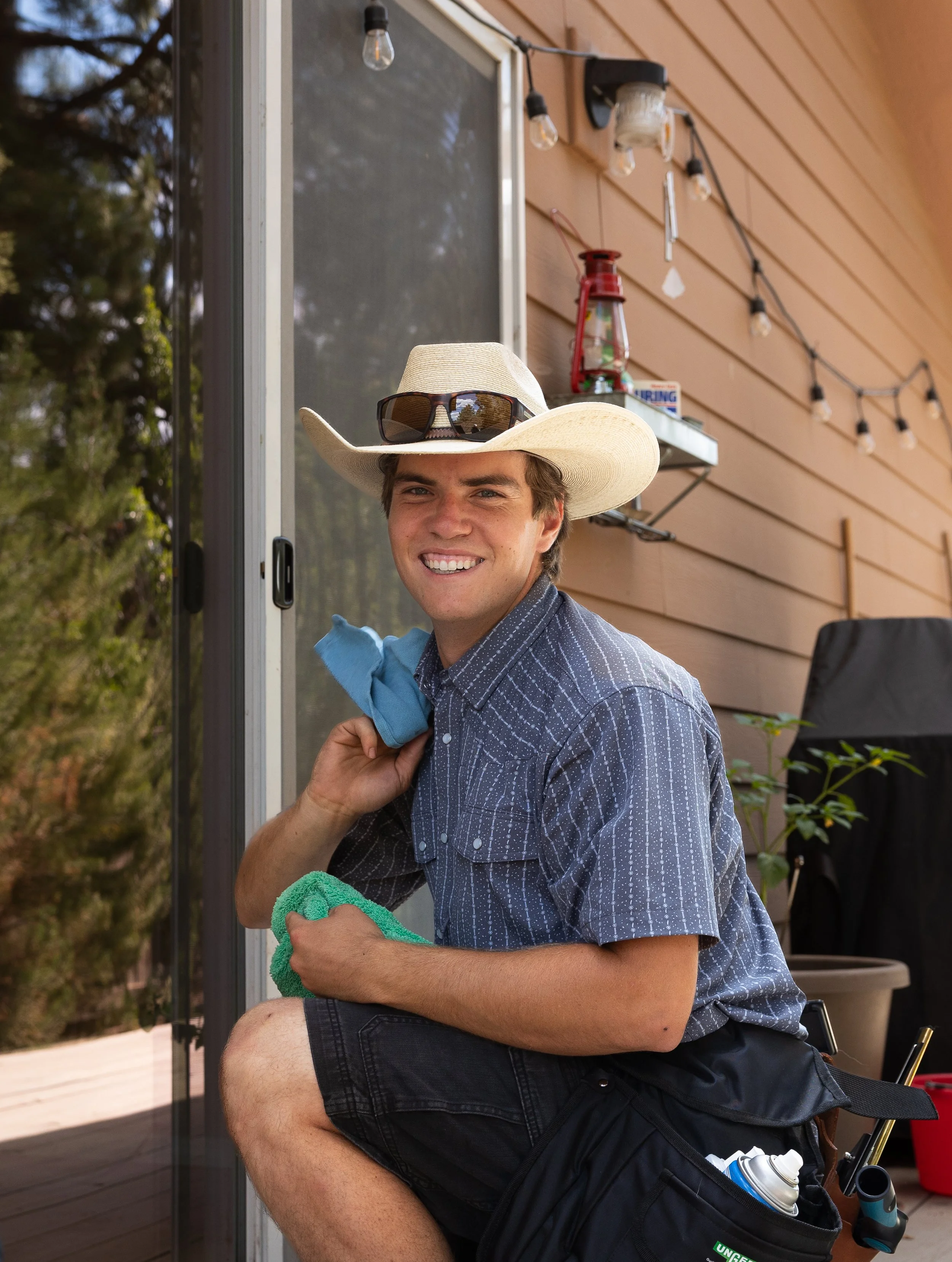 A young man wearing a cowboy hat and sunglasses, smiling while holding cleaning cloths, crouching outside a house with a brown exterior, string lights, a shelf with a lantern and a box, and a potted plant in the background.