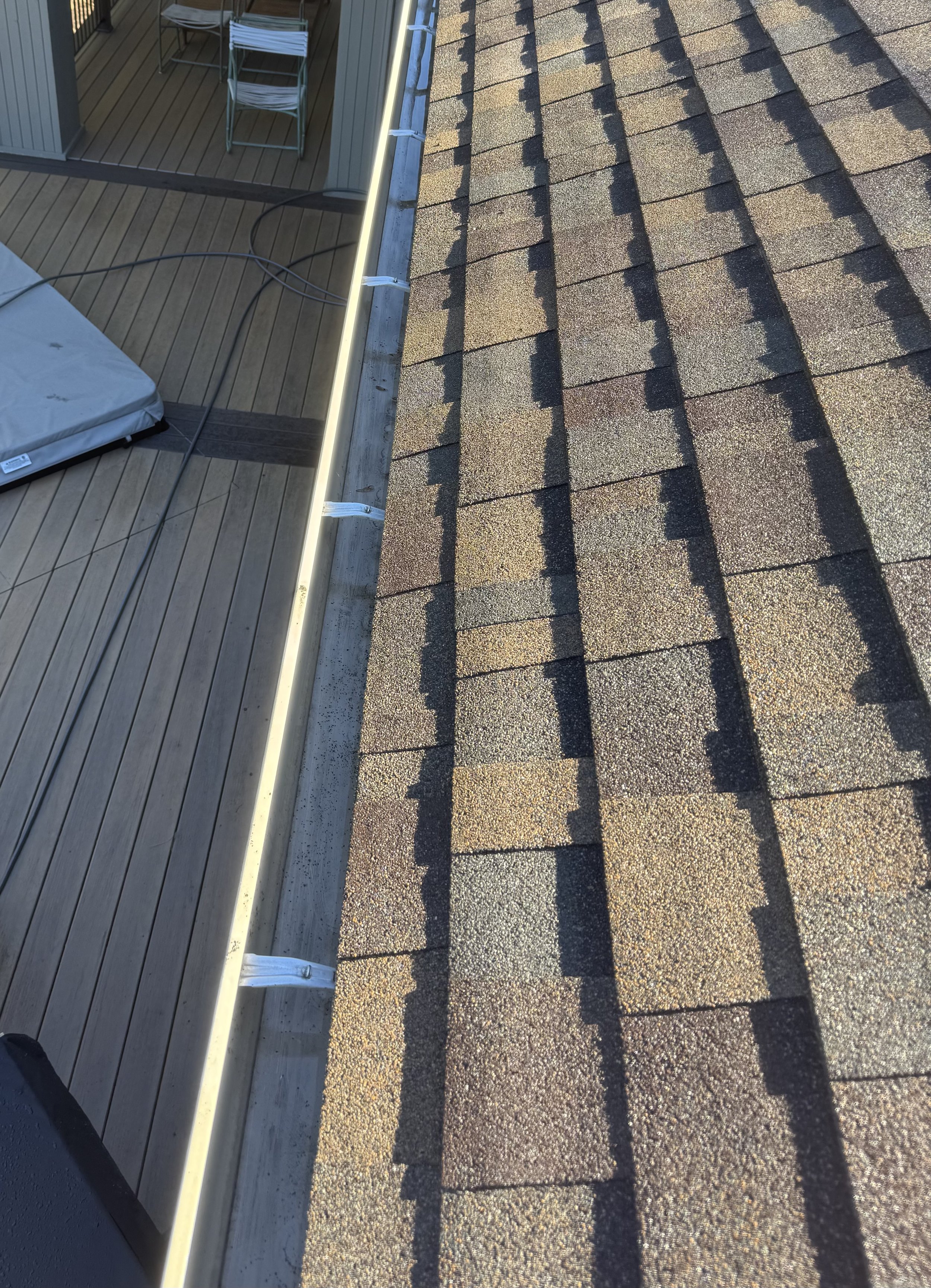 View of a roof with asphalt shingles and a section of a deck with wood planks, metal chairs, a tarp, and some electrical cords, taken from an angled perspective.