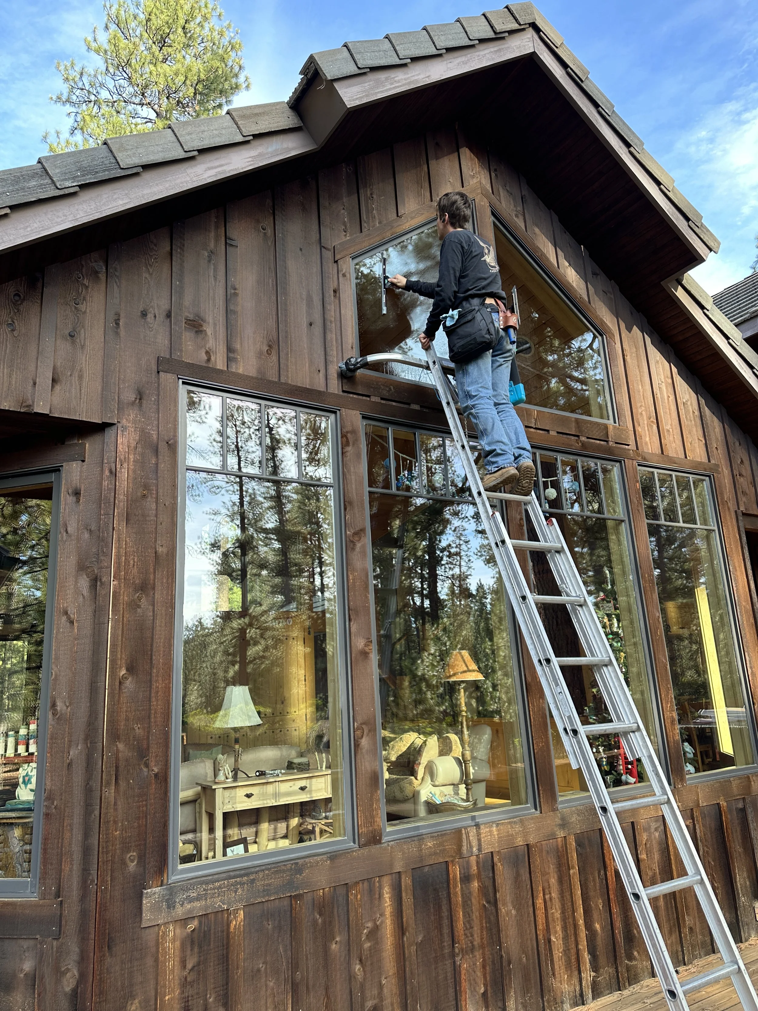 A man on a ladder cleaning or installing a window on a wooden house exterior surrounded by trees and a blue sky.