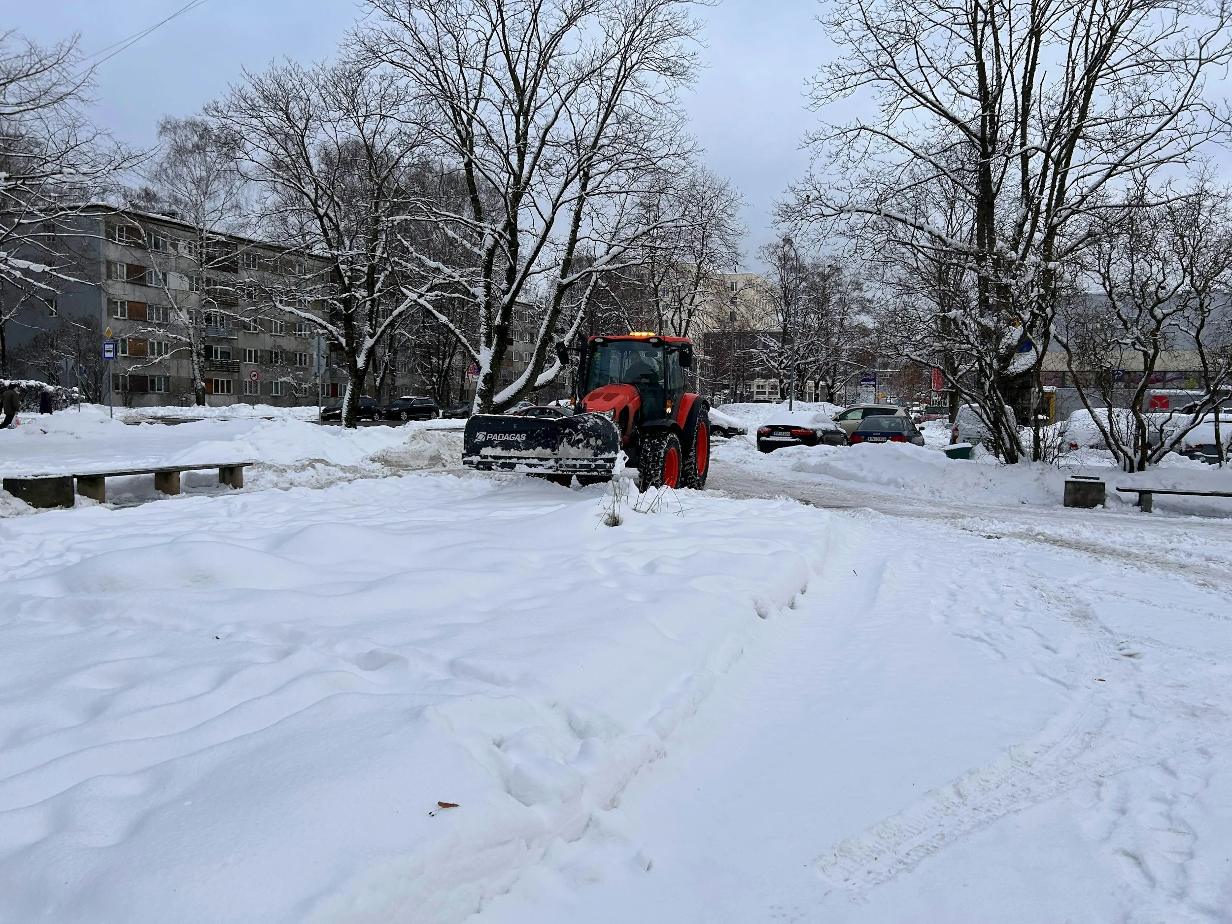 A snow-covered urban park or street with a red snow plow clearing snow, parked cars, trees, and apartment buildings in the background.