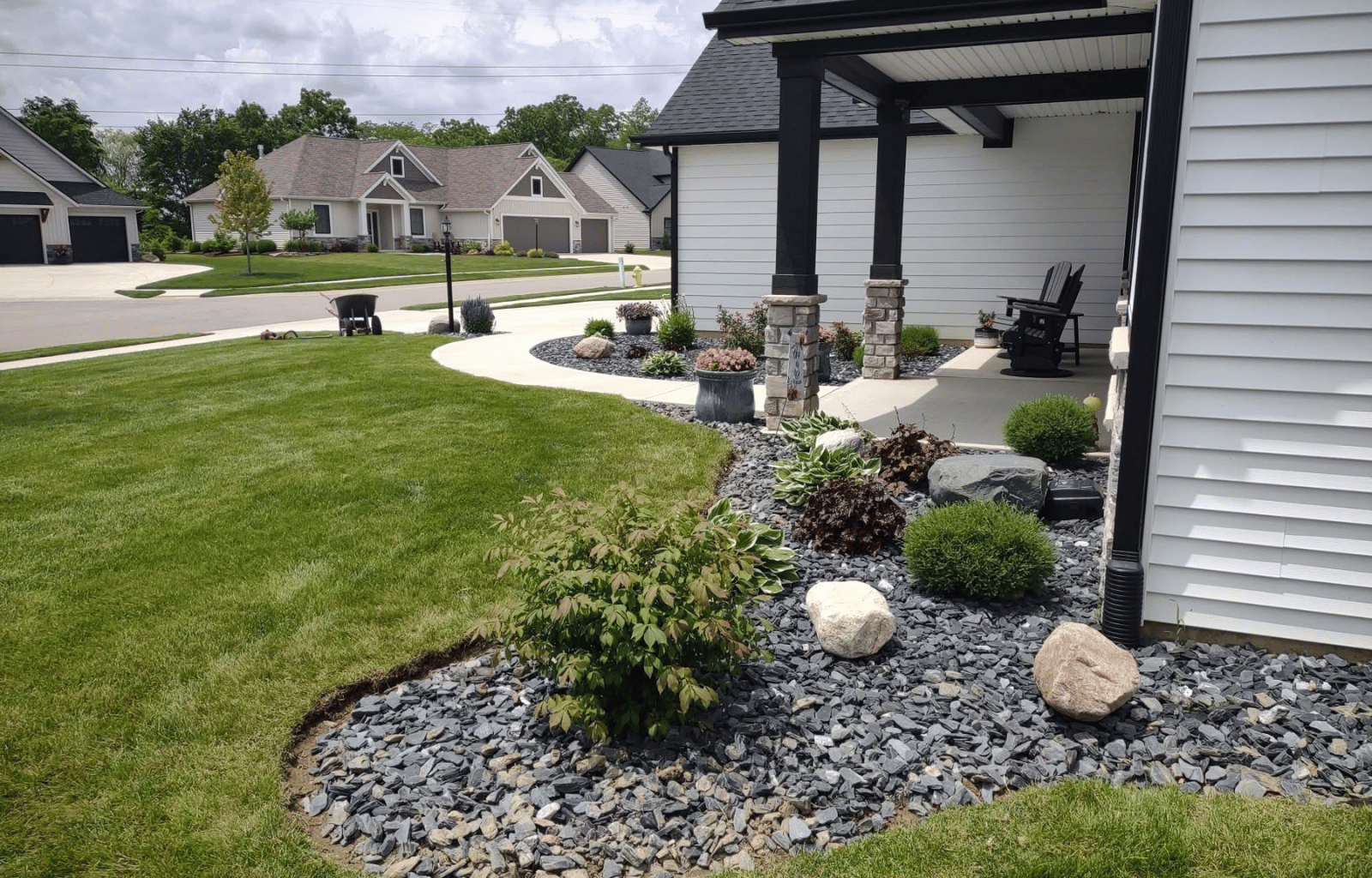 A landscaped front yard with a curved concrete pathway, green grass, shrubs, rocks, and a porch with two black chairs, in a suburban neighborhood with houses in the background.