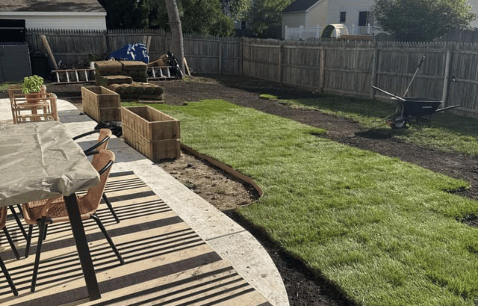 View of a backyard with a patio, outdoor furniture, and a partially finished lawn with garden beds and a wheelbarrow.