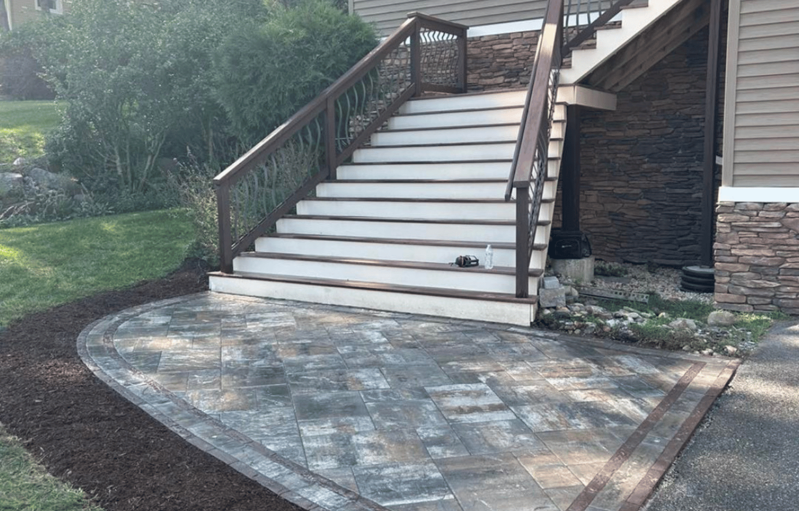 Outdoor patio area with stone paving, adjacent to a set of wooden stairs leading to a house deck. The background includes greenery and shrubs.