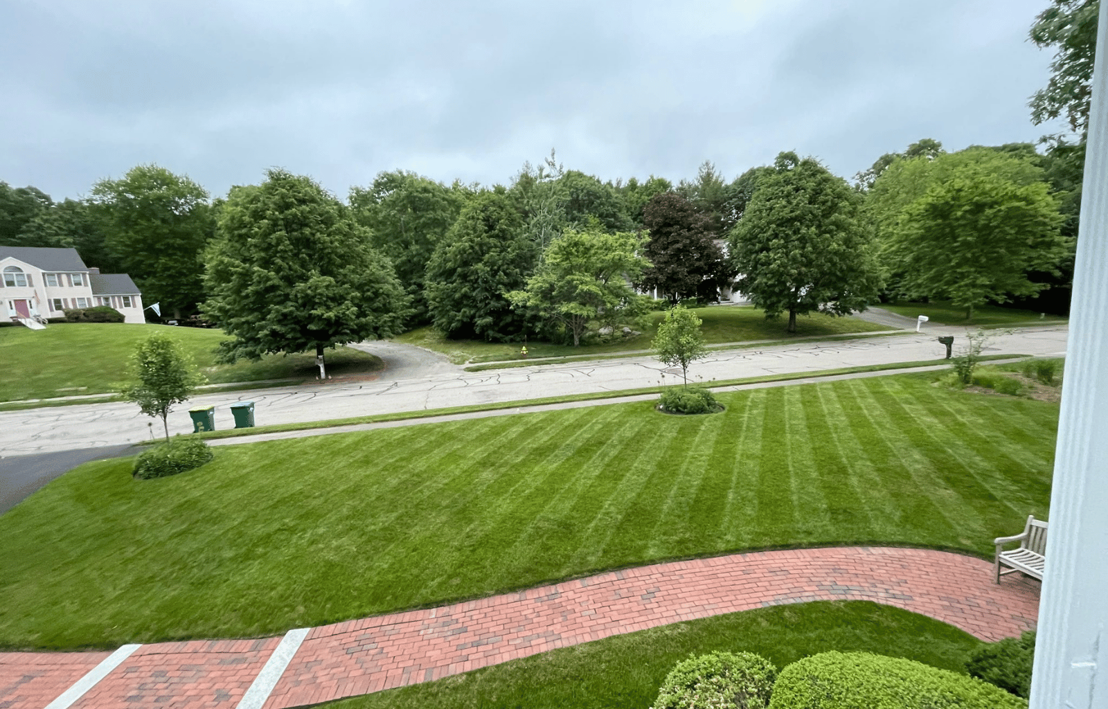 View of a suburban neighborhood street with trees, houses, a grassy yard, a brick walkway, and two green trash bins on a cloudy day.