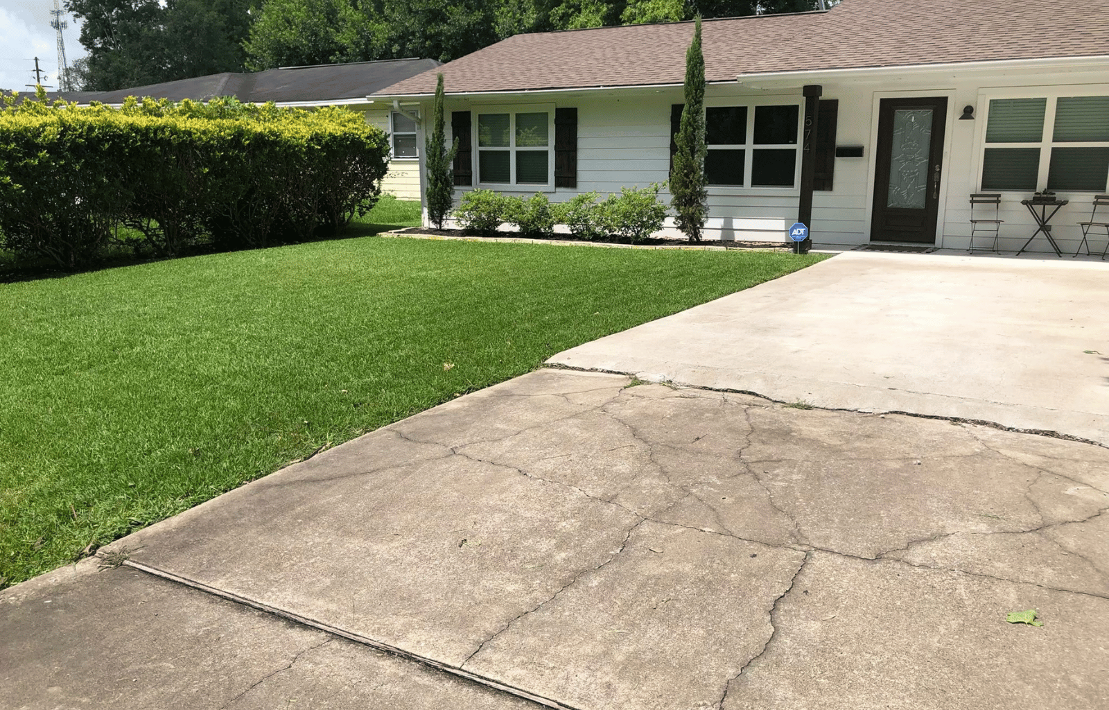 Front yard of a house with a well-maintained lawn, small bushes, two tall thin trees, a driveway with visible cracks, and a white house with black shutters and a porch with metal chairs and a table.