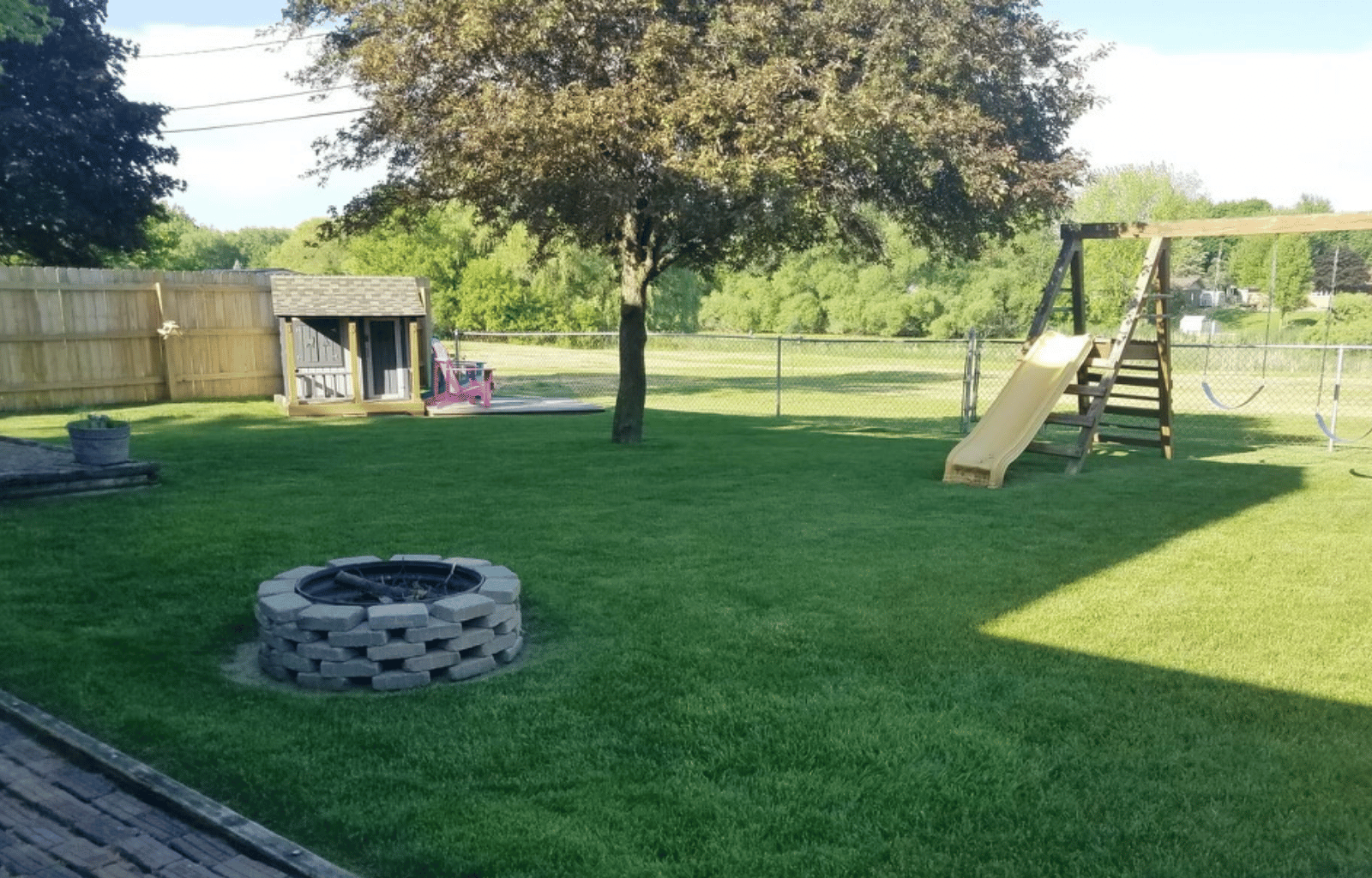 Backyard with green lawn, trees, a wooden swing set with slide, a fire pit made of bricks, a wooden shed, and a fence.