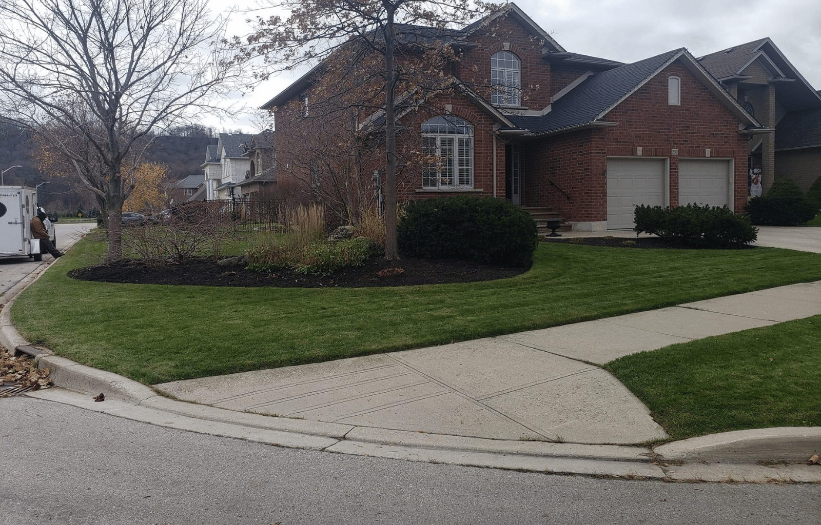 A suburban house with red brick exterior, split garage doors, and a well-maintained front yard with mulch, bushes, and a small tree, street corner view on a cloudy day.