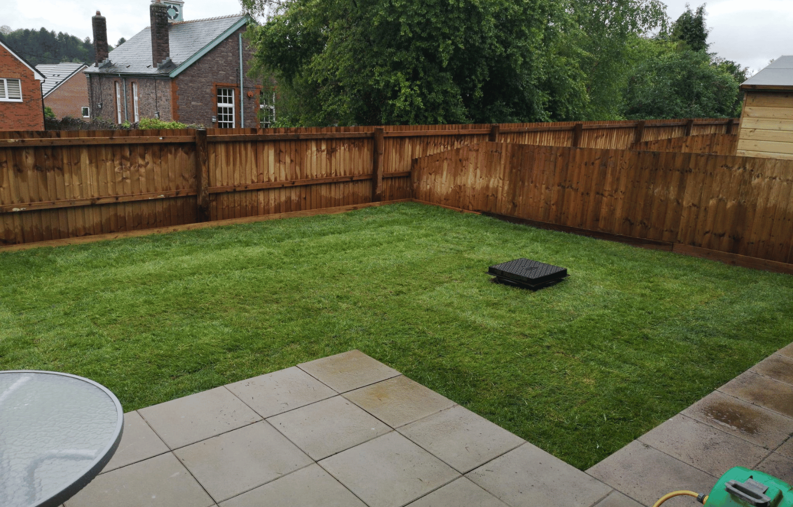 Empty backyard with green grass, wooden fence, and neighboring houses in the background, partially showing a patio with tiled flooring and a round glass table.