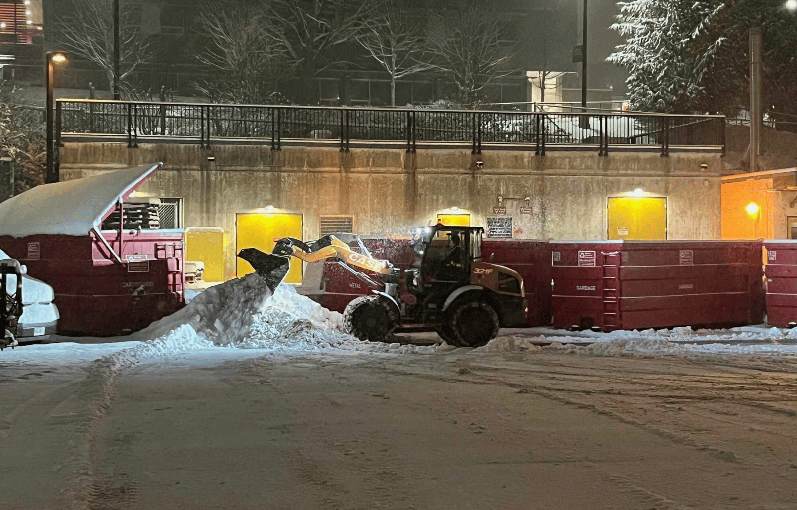 A snow-covered urban parking lot at night with a snowplow clearing snow, surrounded by red garbage bins, a truck, and a building with yellow-lit doors, snow-covered trees, and a fence on the elevated background.