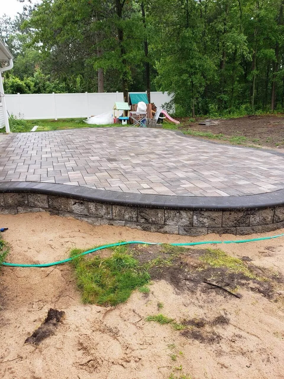 Newly paved patio with brick pavers and stone border in a backyard, with children's play equipment and a fenced yard in the background, and patchy dirt and grass in the foreground.