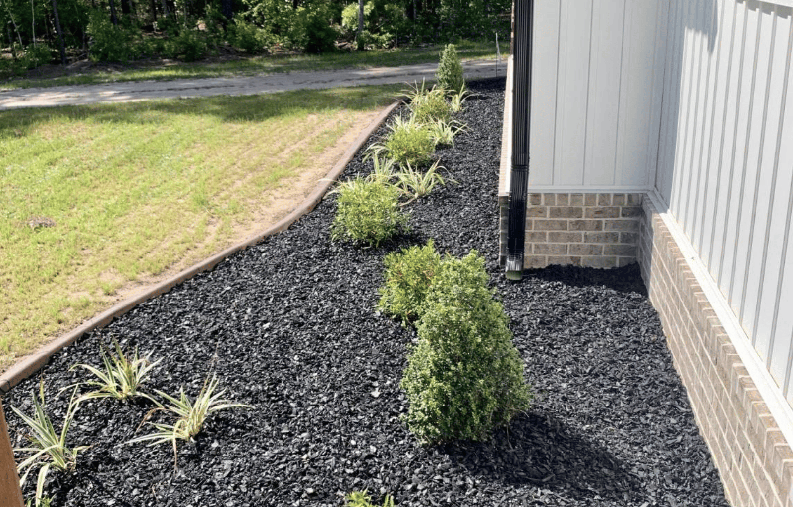 A landscaped yard with a mulch garden bed next to a house with brick and siding exterior. The garden contains small bushes and plants, bordered by a wooden edge, with a grassy lawn and trees in the background.