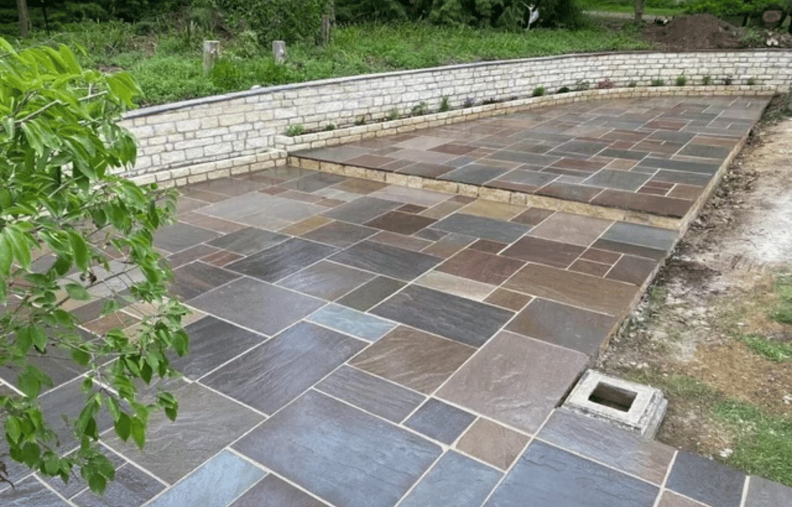 Outdoor patio with multicolored stone tiles, bordered by a curved white brick wall, and surrounded by greenery and garden beds.