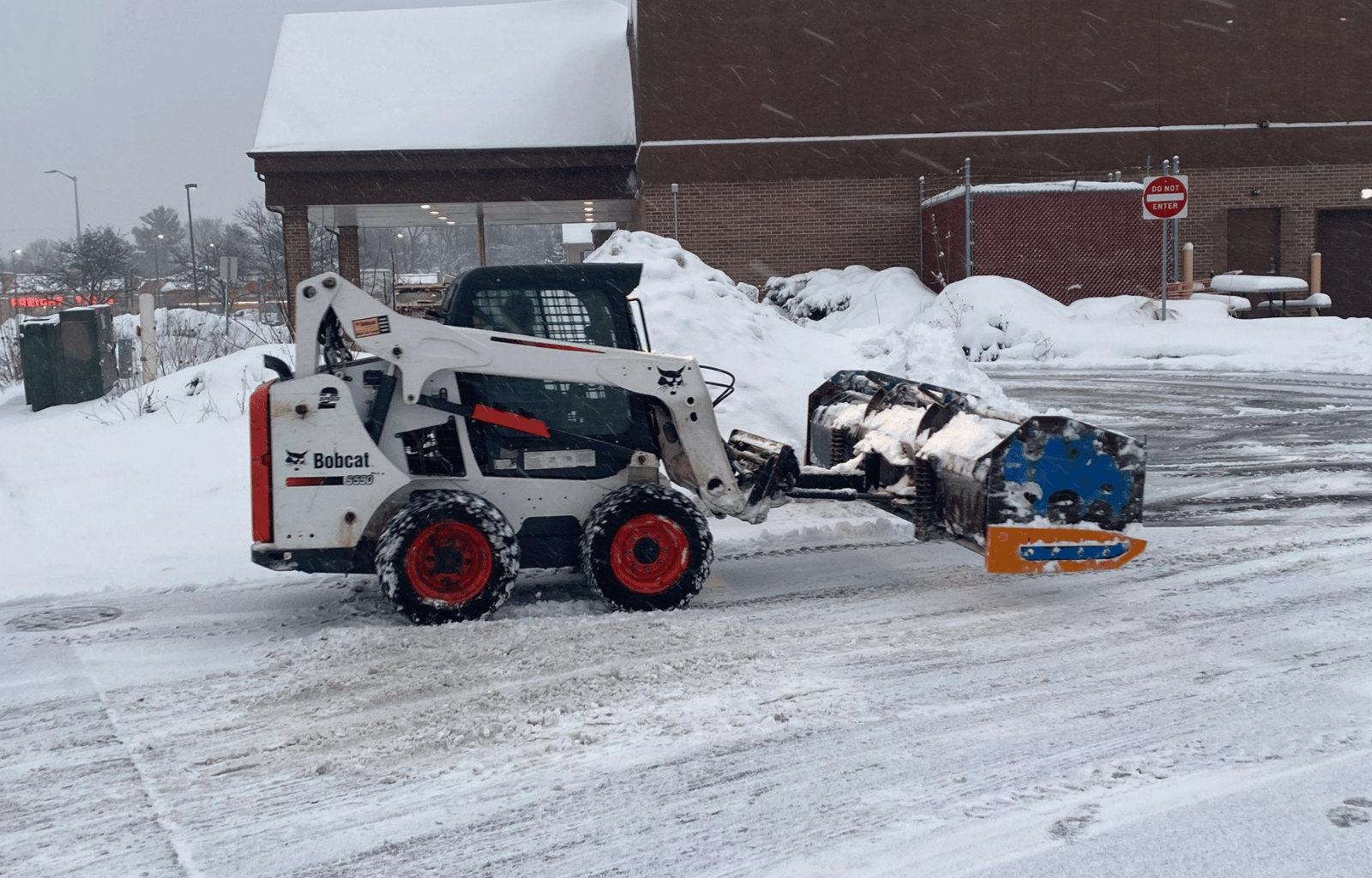 A Bobcat skid-steer loader clearing snow from a parking lot during a snowy day.