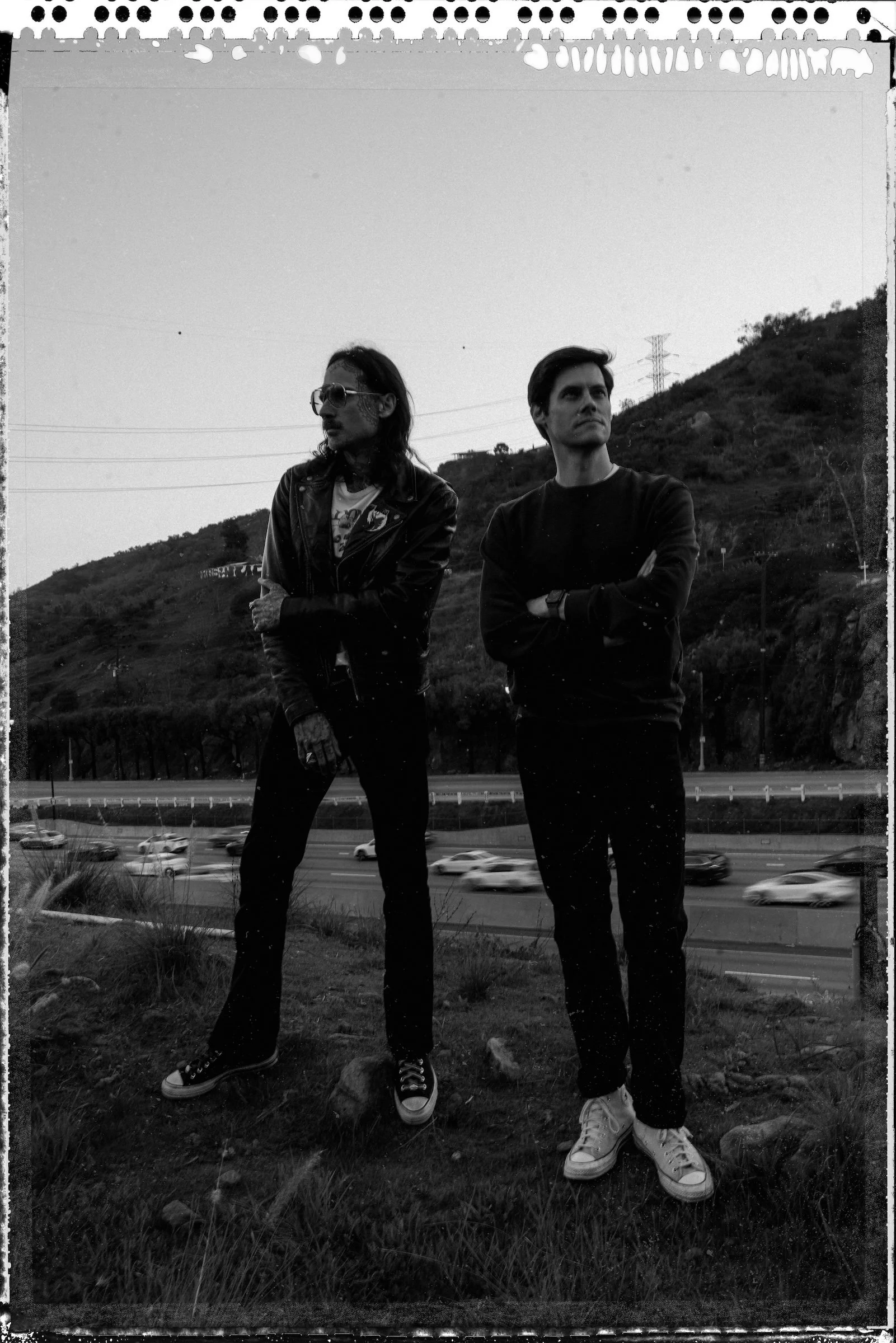 KaL MichaeL and Art Quanstrom.Two men standing outdoors near a highway with hills in the background, both with arms crossed, one wearing sunglasses, leather jacket, and sneakers, the other in a dark sweatshirt and white sneakers, during dusk or dawn.
