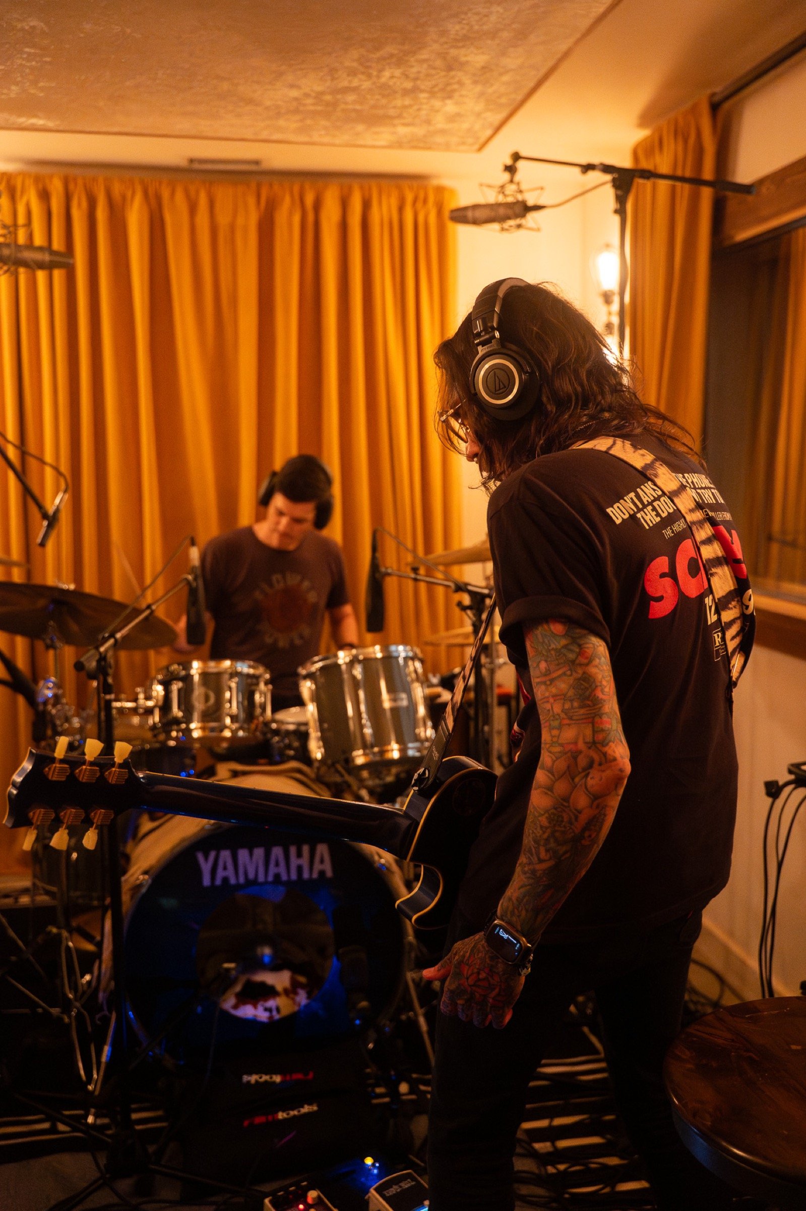Two musicians in a recording studio; one playing an electric guitar, the other on drums. Both wear headphones, the guitarist is focused on his instrument, and the drummer is in the background. The room has warm, yellow curtains.