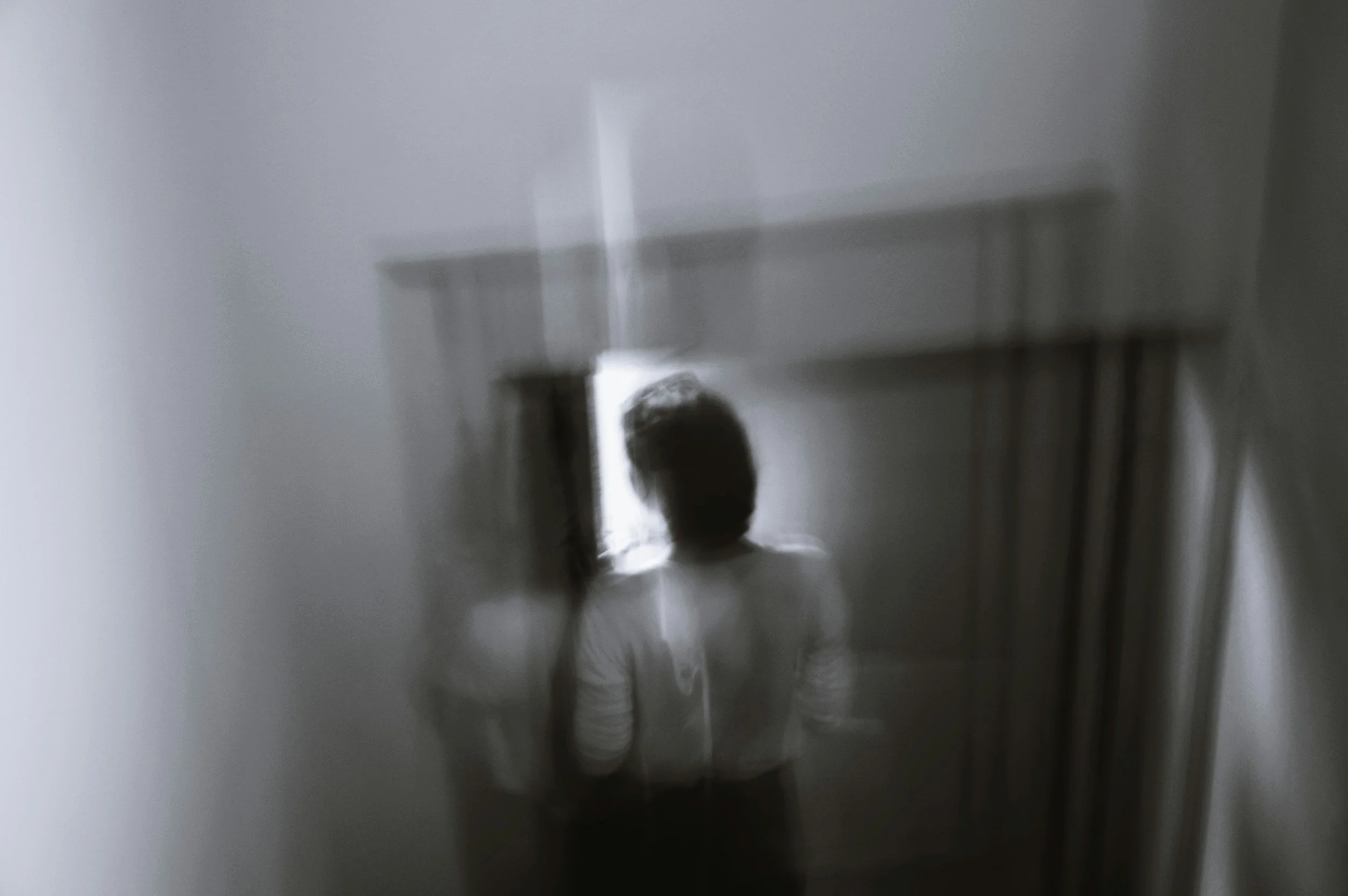 A person standing in front of an illuminated doorway, seen through a semi-transparent window or blurred glass.