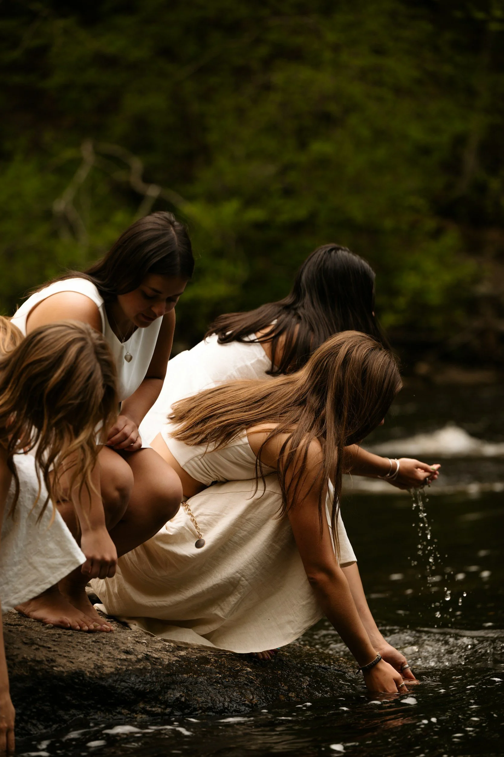 Group of young women wearing light-colored dresses crouching by a river, dipping their hands into the water, surrounded by green trees.