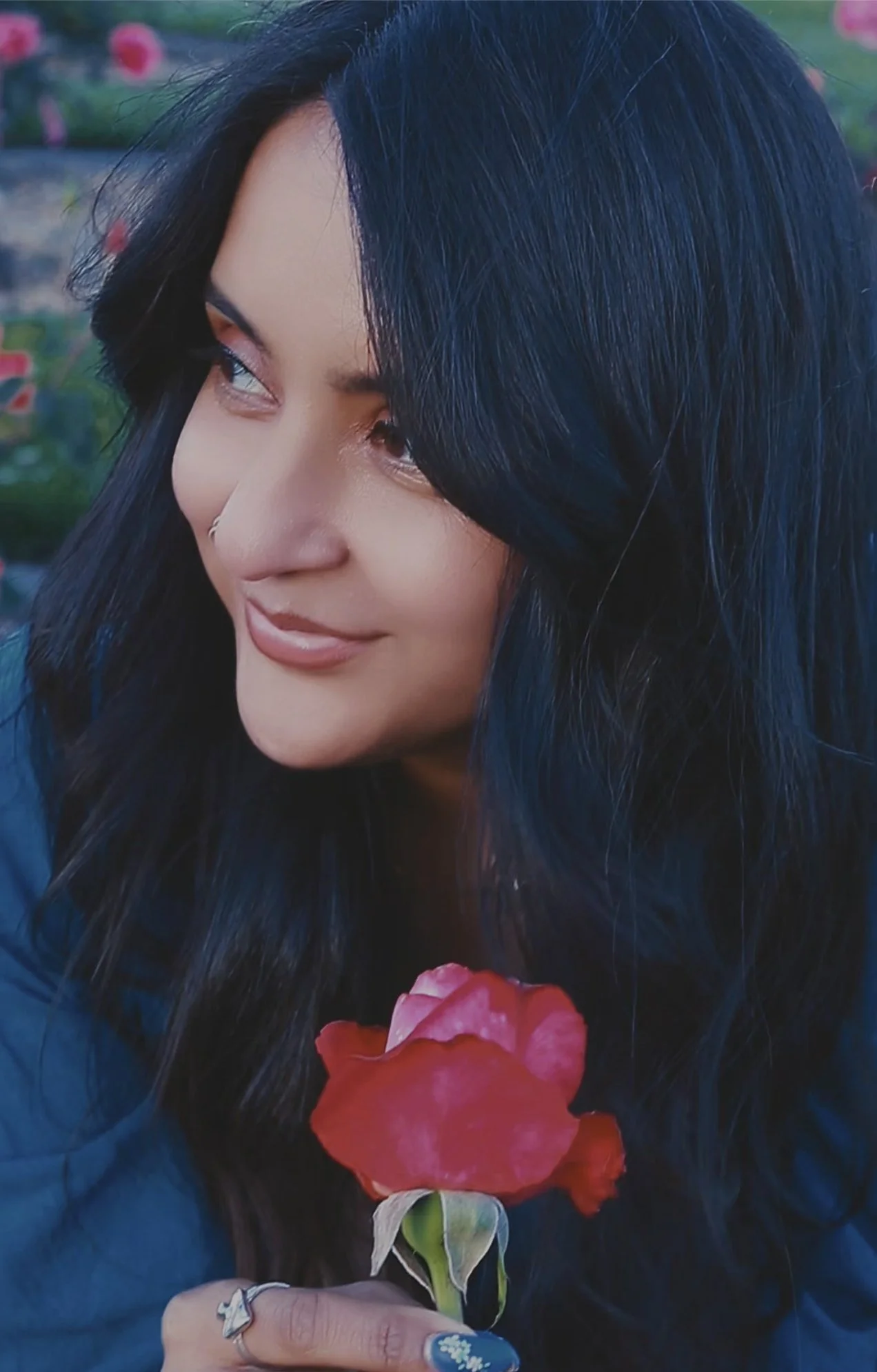 A woman with long black hair and light makeup holds a red rose, gazing to the side with a gentle smile, outdoors with pink flowers in the background.