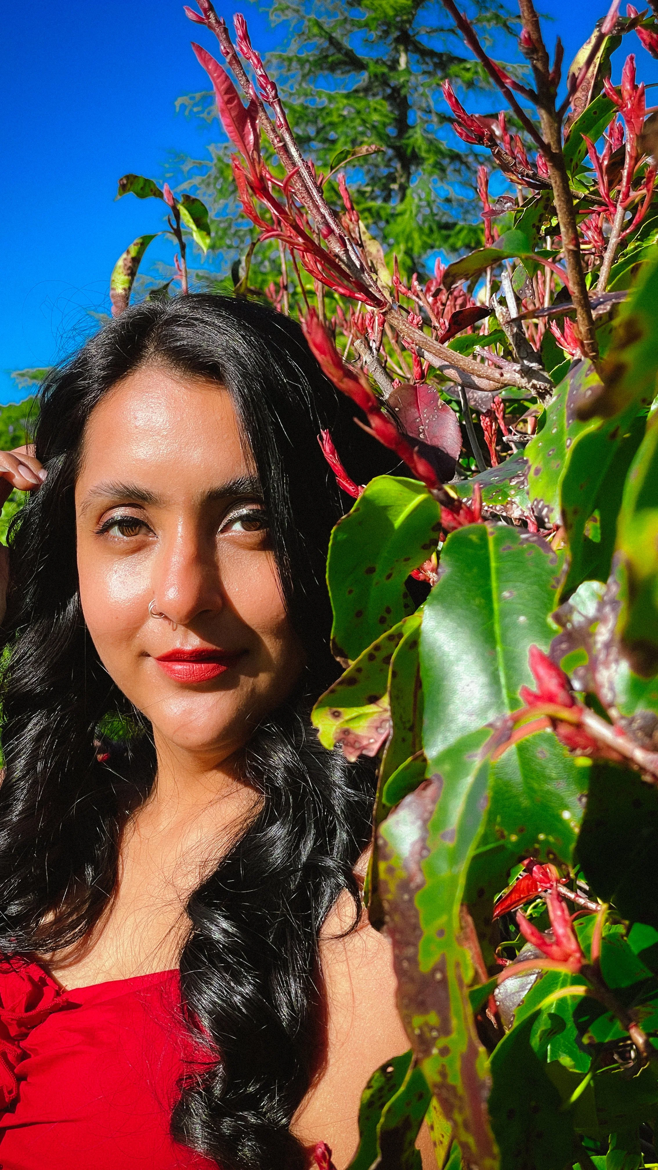 A woman with long black curly hair, wearing a red top, is taking a close-up selfie outdoors. She is partly obscured by green and reddish leaves, with a bright blue sky in the background.