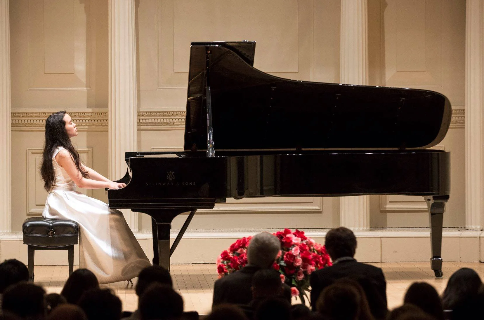 A young woman in a white dress playing a grand piano on stage in front of an audience.