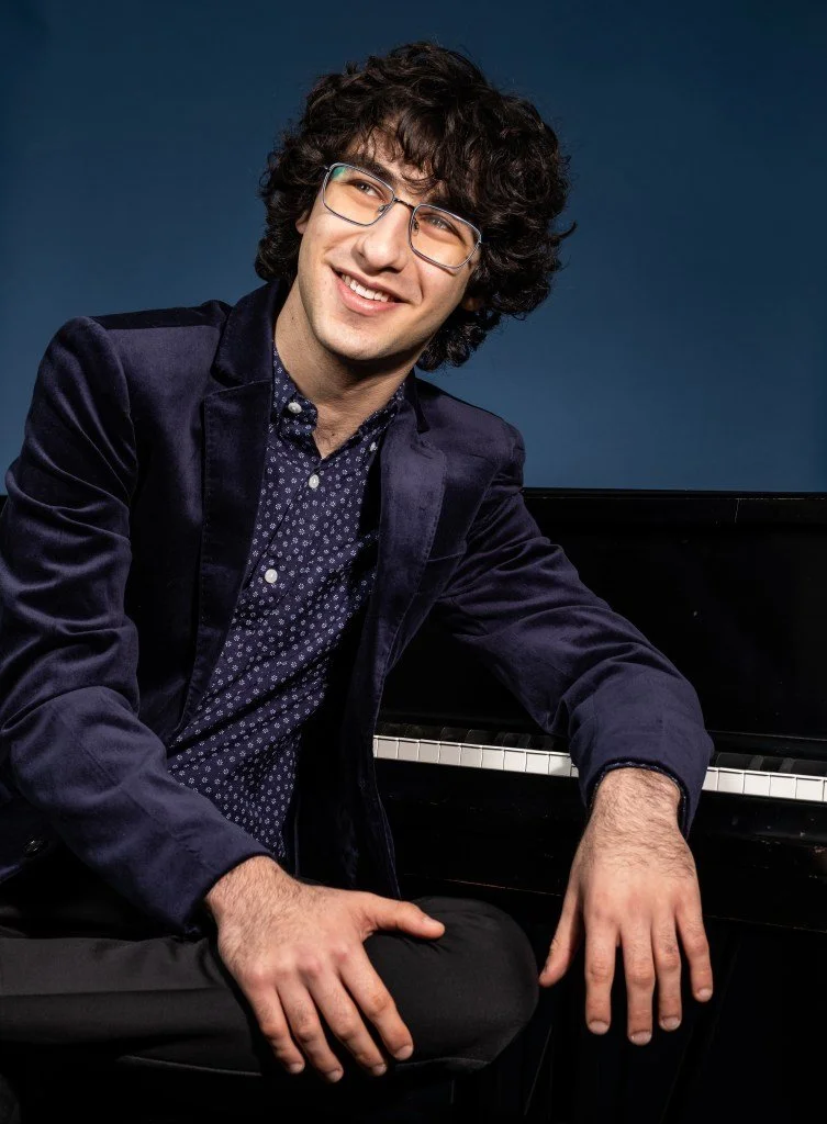 A young man with curly dark hair and glasses, sitting next to a black piano, smiling in a portrait against a dark blue background.