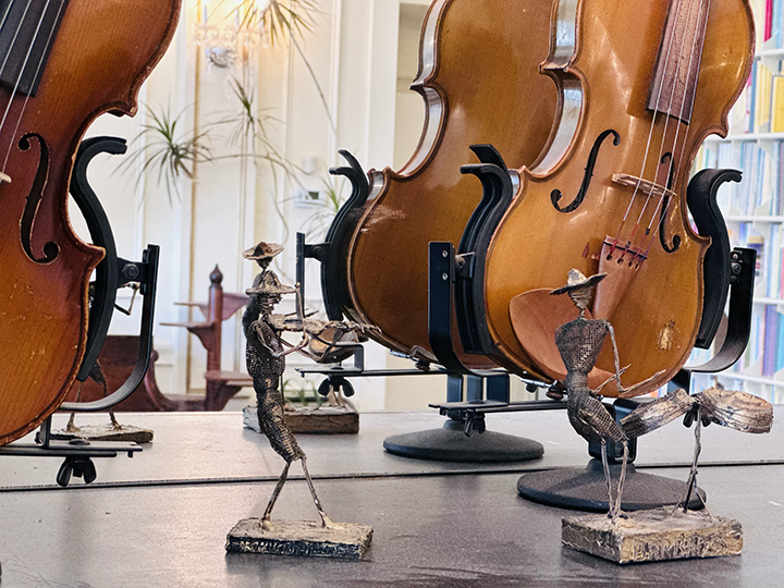 Three sketches of musicians playing violins in front of real violins on stands, with bookshelves in the background.