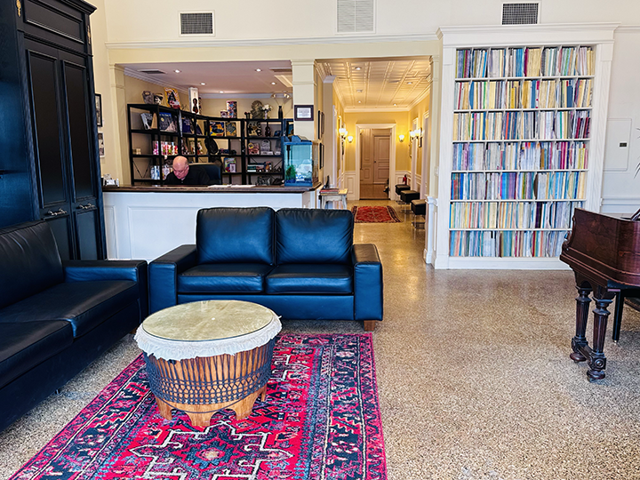 Living room with black leather sofas, a colorful patterned rug, a round drum-shaped ottoman, a tall bookshelf filled with books, a piano, and a person at the reception desk in the background.