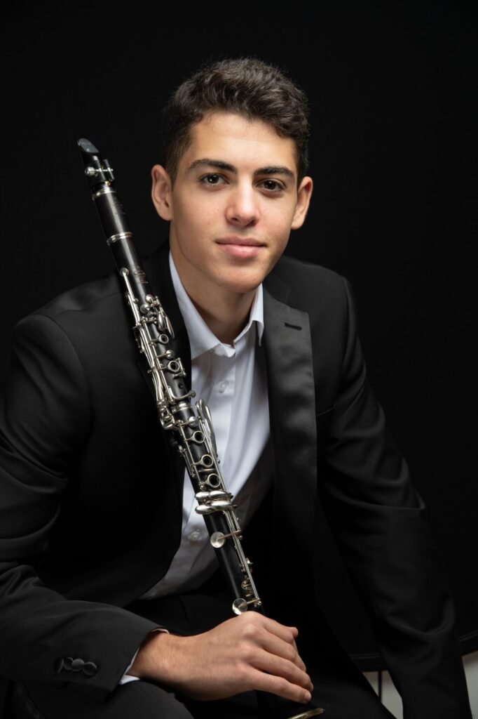 Young man in tuxedo holding a clarinet against a black background.
