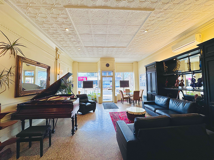 Interior of a spacious lounge with a grand piano, black leather sofas, a large mirror, and windows with street view