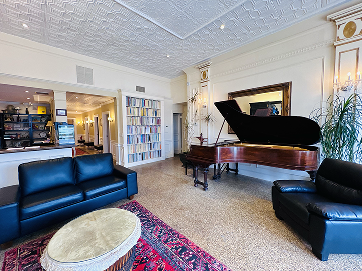 Living room with a grand piano, two black leather sofas, a colorful rug, a bookshelf filled with books, and potted plants.