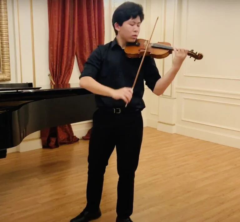 A young man playing a violin in a formal room with wooden floors, cream-colored walls, red curtains, and a black grand piano.
