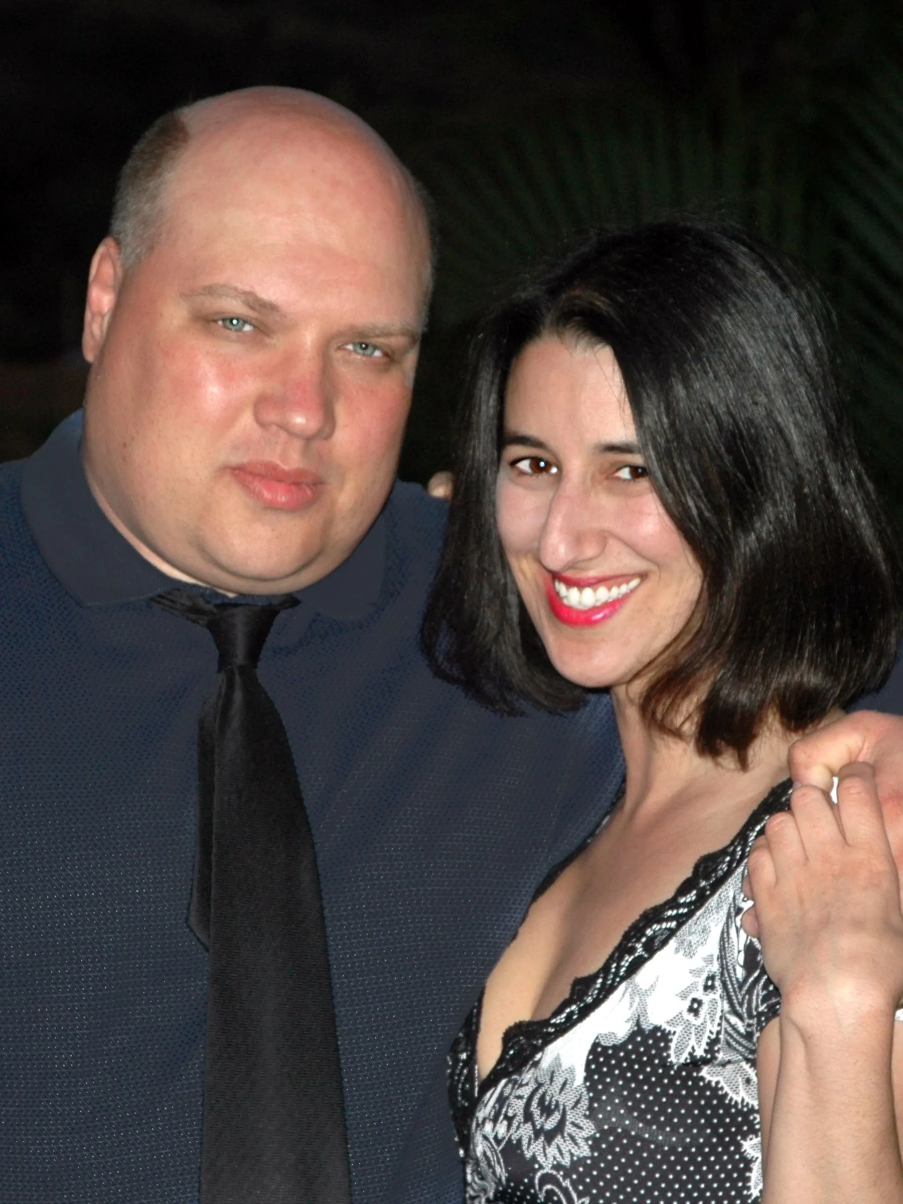 A smiling woman with black hair and red lipstick standing close to a man with light skin, blue eyes, wearing a black suit and tie, in a dark setting.
