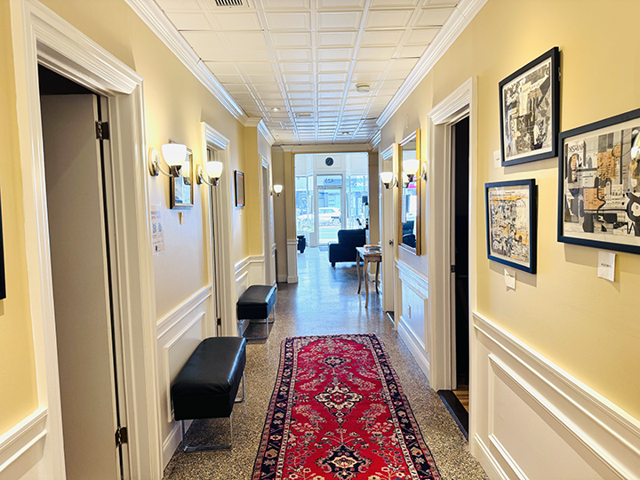 Interior hallway with yellow walls, framed artwork, black benches, and a red patterned rug leading to a sitting area with sofas and large windows.