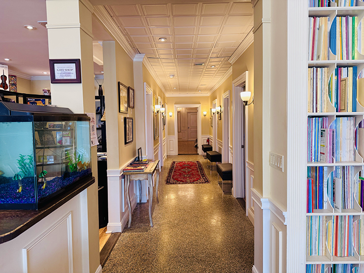 A hallway in a building decorated with framed pictures, wall sconces, and a red rug running along the floor, with seats on the right side and a door at the end.