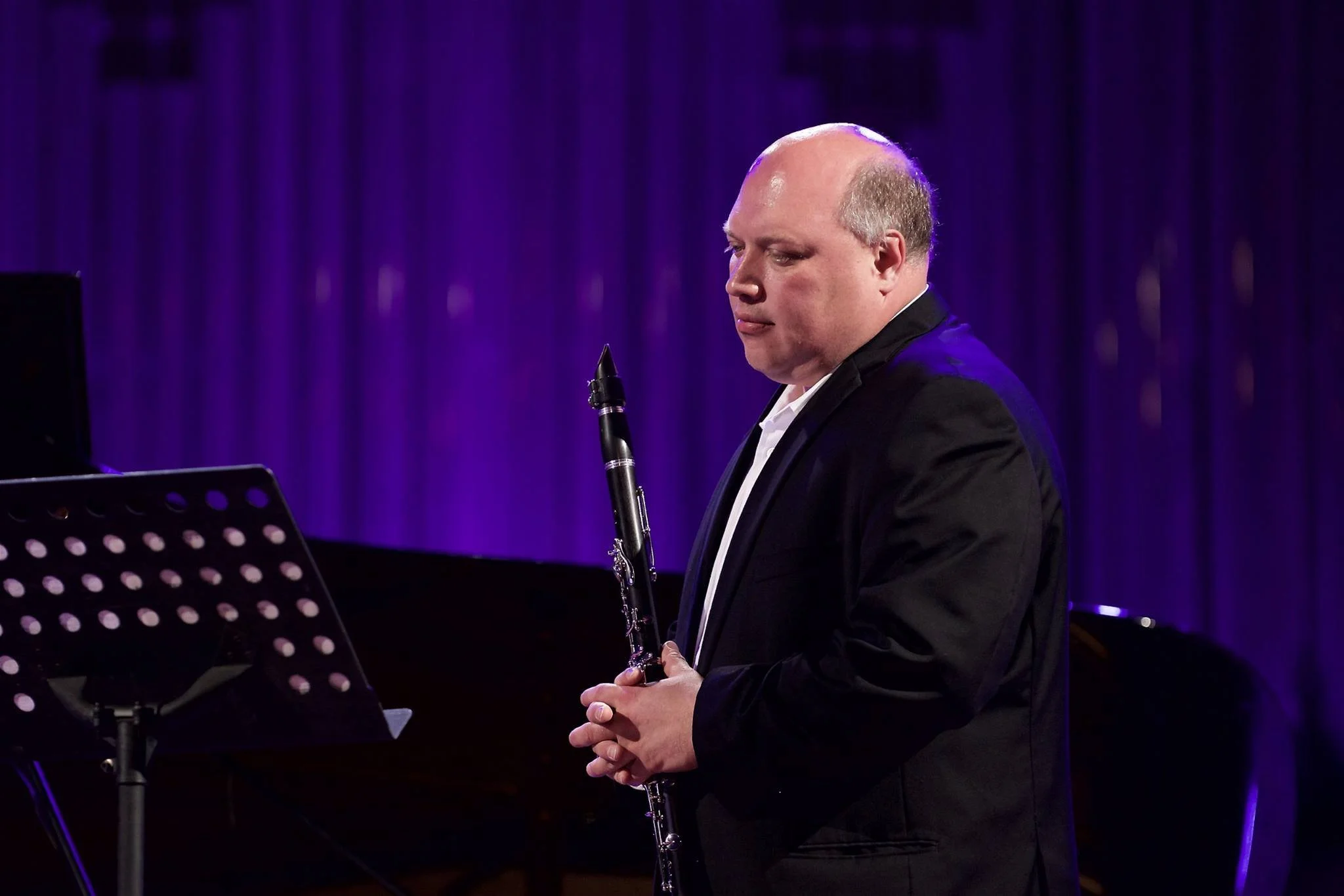 A man with a bald head and light skin wearing a black suit jacket and white shirt playing a clarinet on a stage with purple curtains in the background.