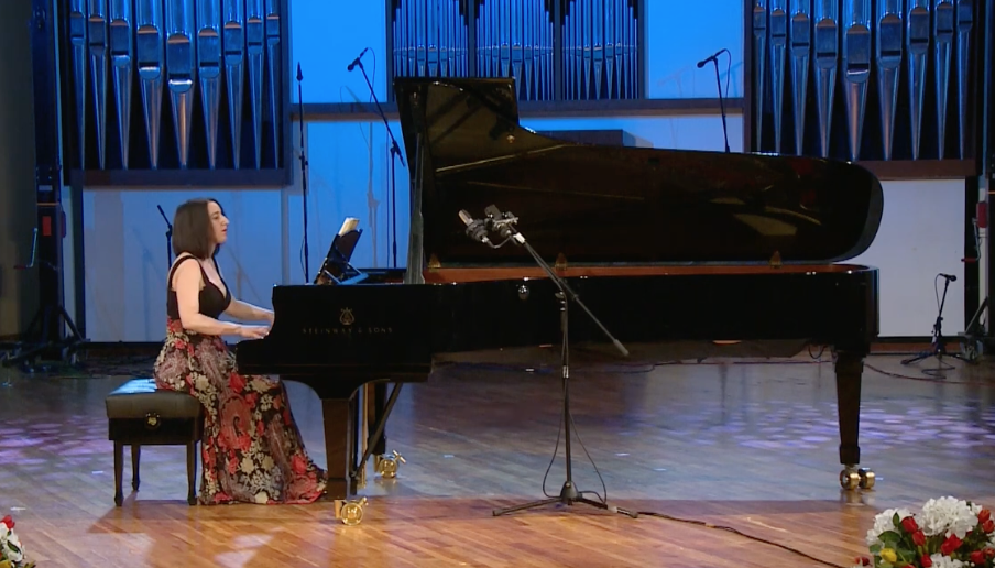 A woman playing a grand piano on a stage with a blue background and microphones, with flowers visible in the lower right corner.